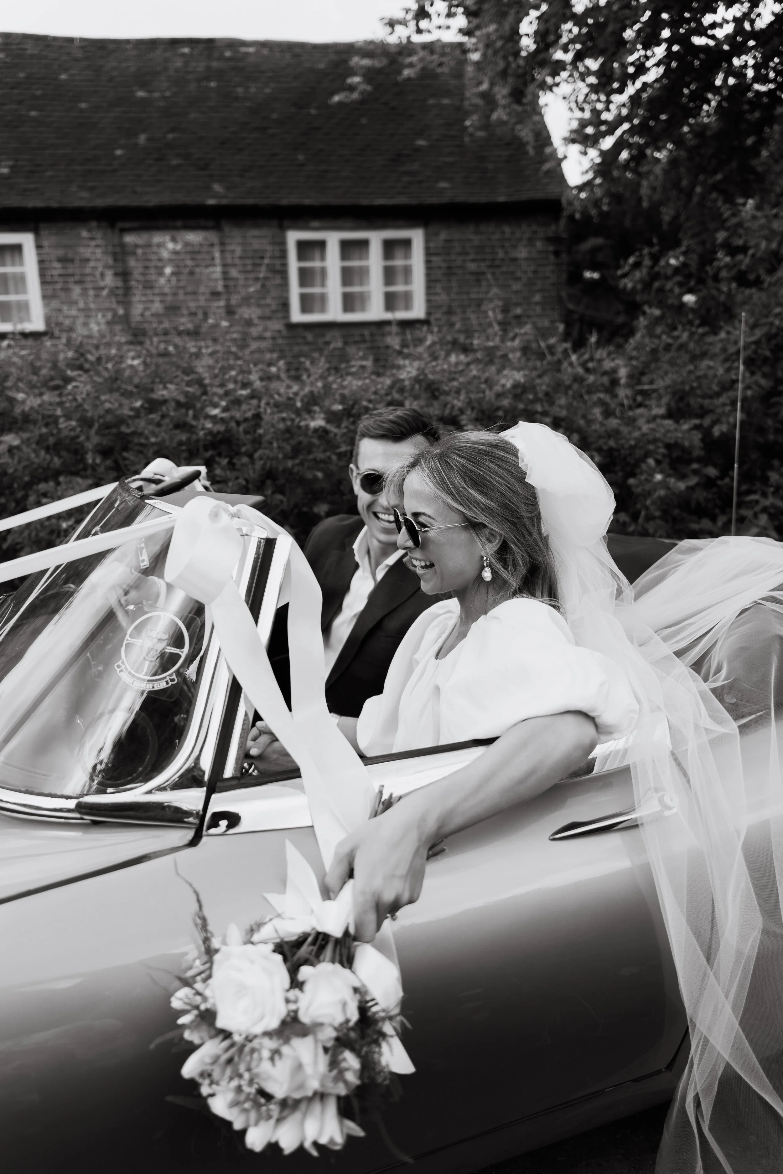 A black-and-white photo of a bride and groom sitting in a vintage convertible car decorated for a wedding, smiling and wearing sunglasses, with a bouquet of flowers in the bride's hand.