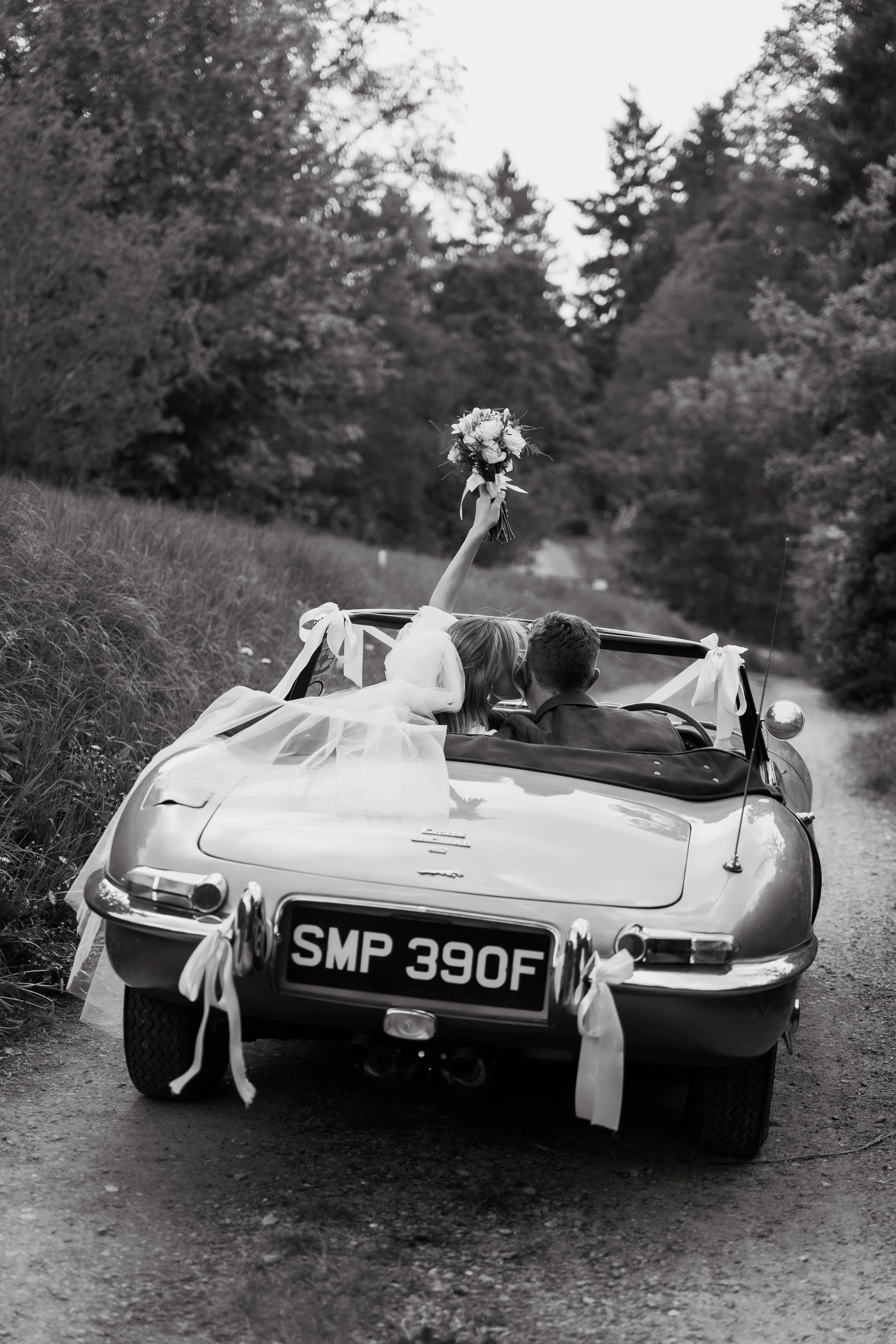 A bride and groom driving in a vintage convertible car decorated with ribbons on a rural dirt road, with the bride holding a bouquet and leaning towards the groom.