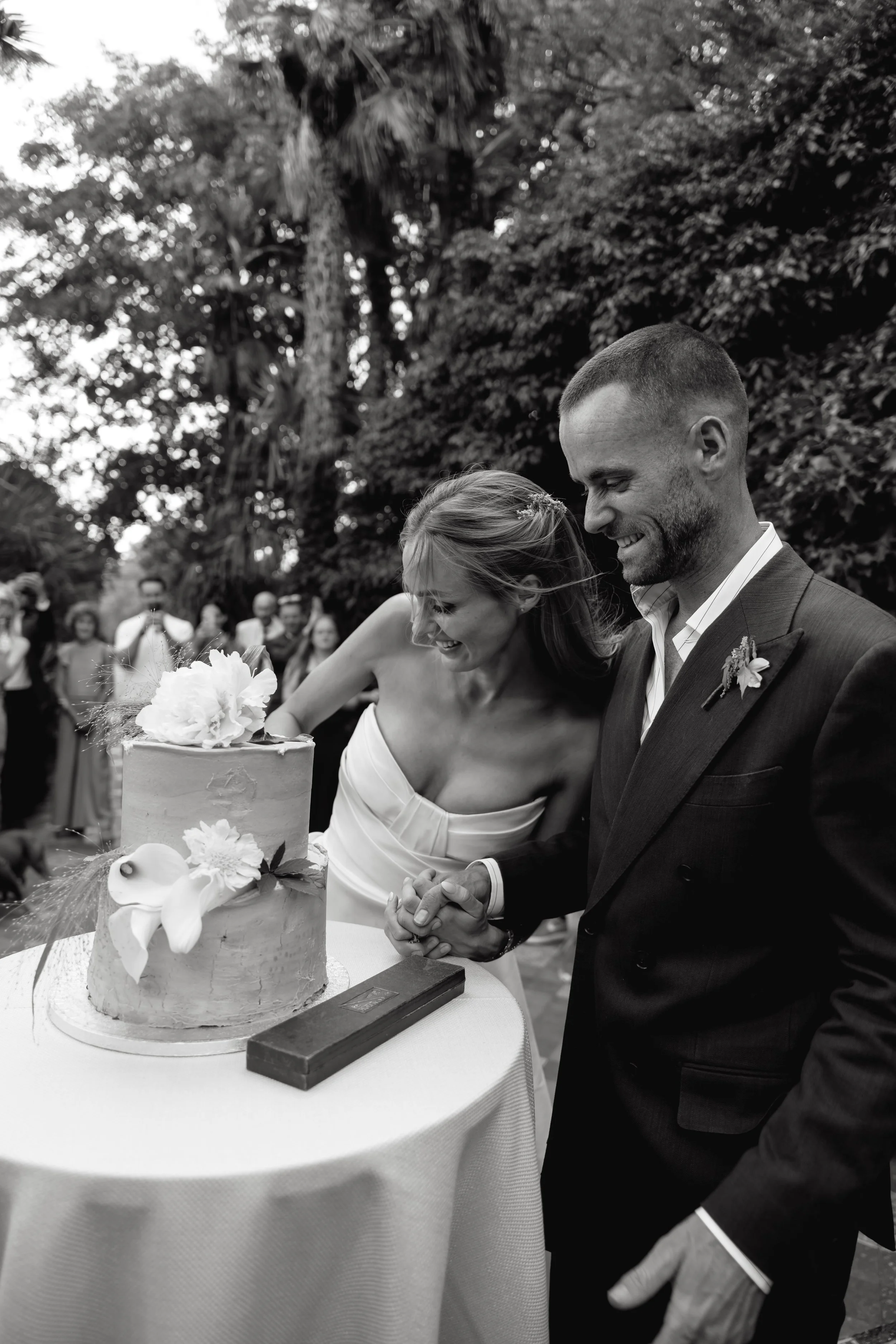 A bride and groom cutting a wedding cake during an outdoor wedding celebration, surrounded by wedding guests, with trees in the background.