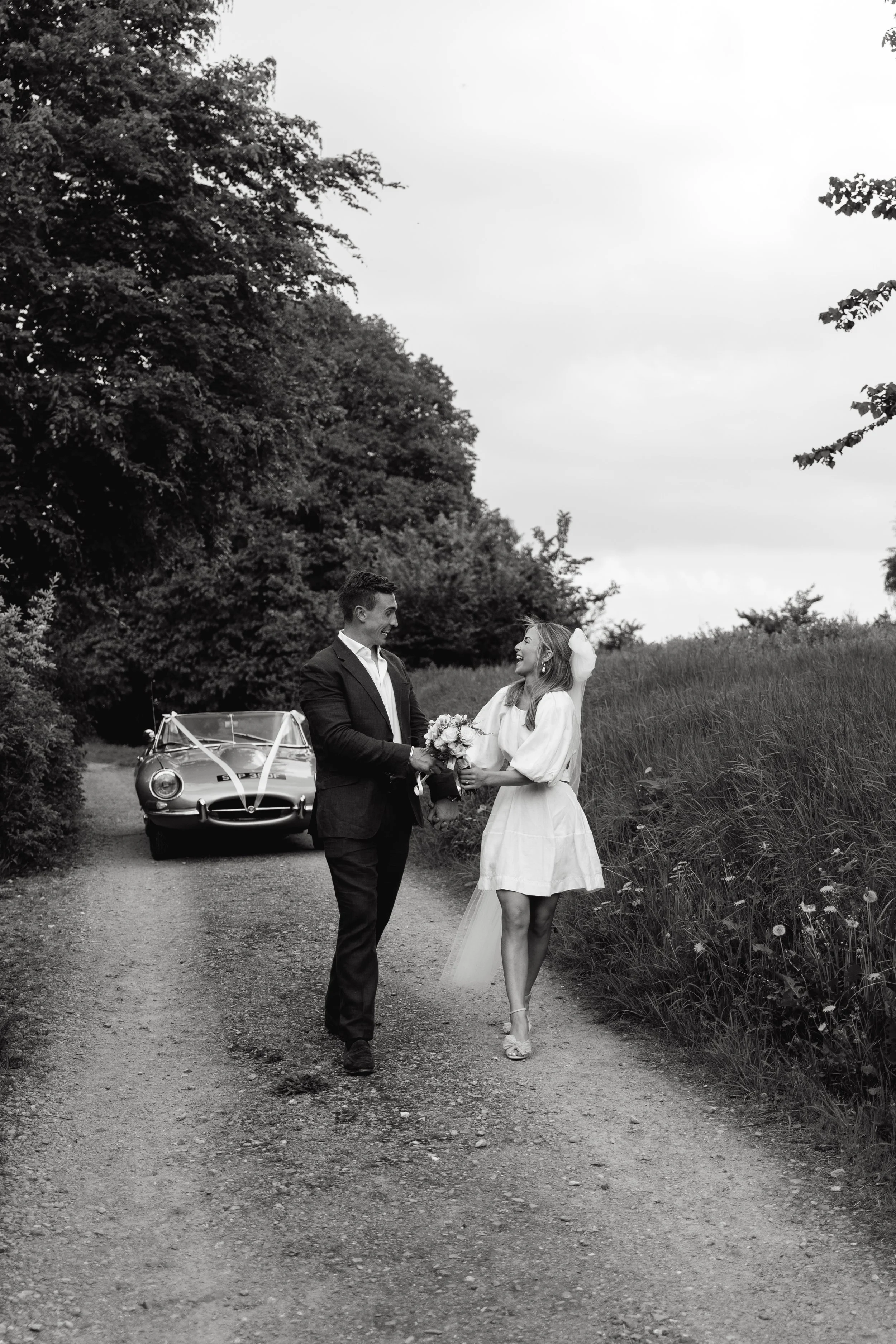 A black and white photo of a couple on a rural path, with a vintage car decorated with ribbons in the background. The man is in a suit offering a bouquet to the woman in a white dress, who is smiling and wearing heels, amidst tall grass and trees.