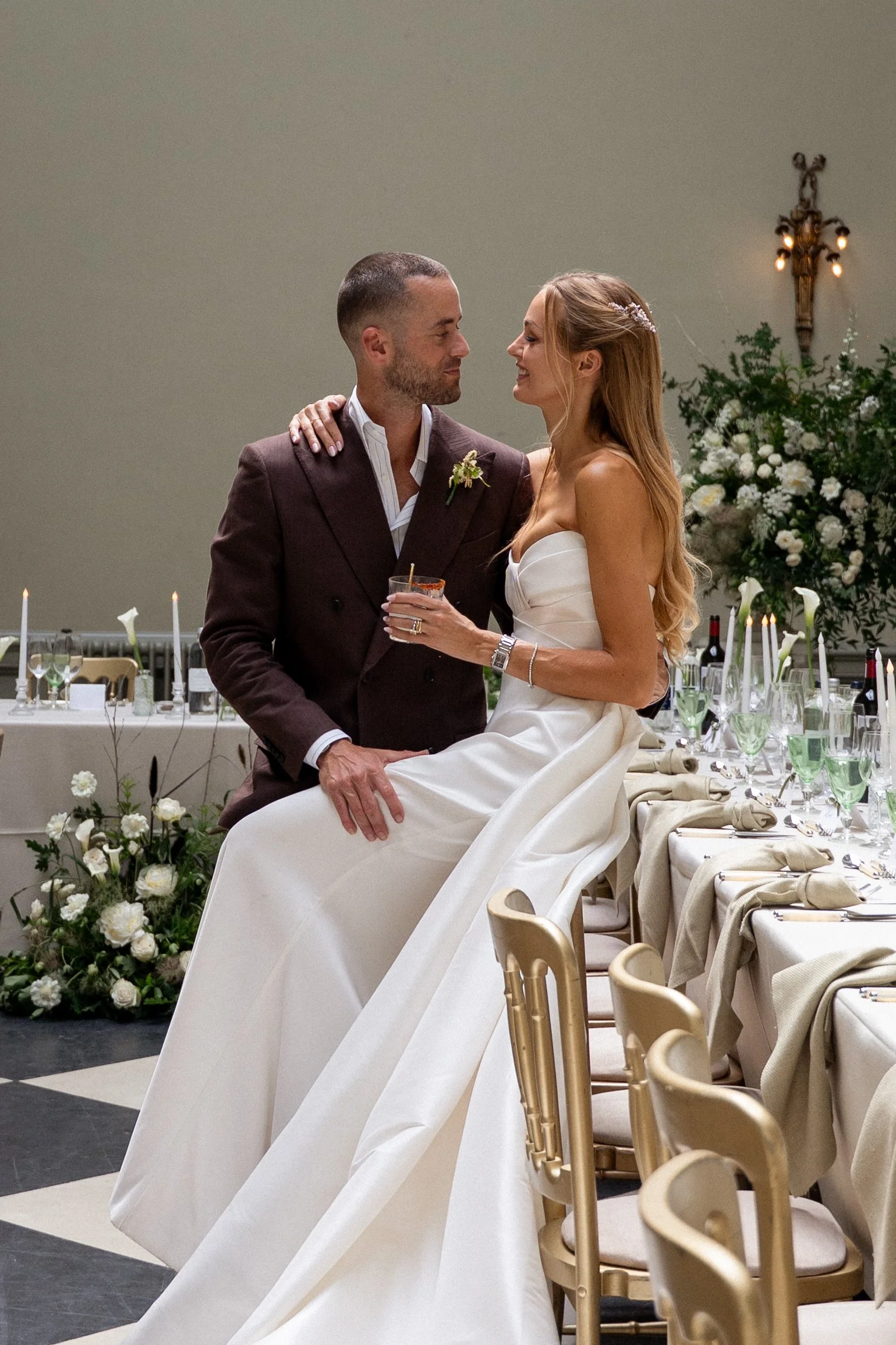 A bride and groom sharing a laugh at their wedding reception, sitting on a table with elegant decorations and floral arrangements in the background.