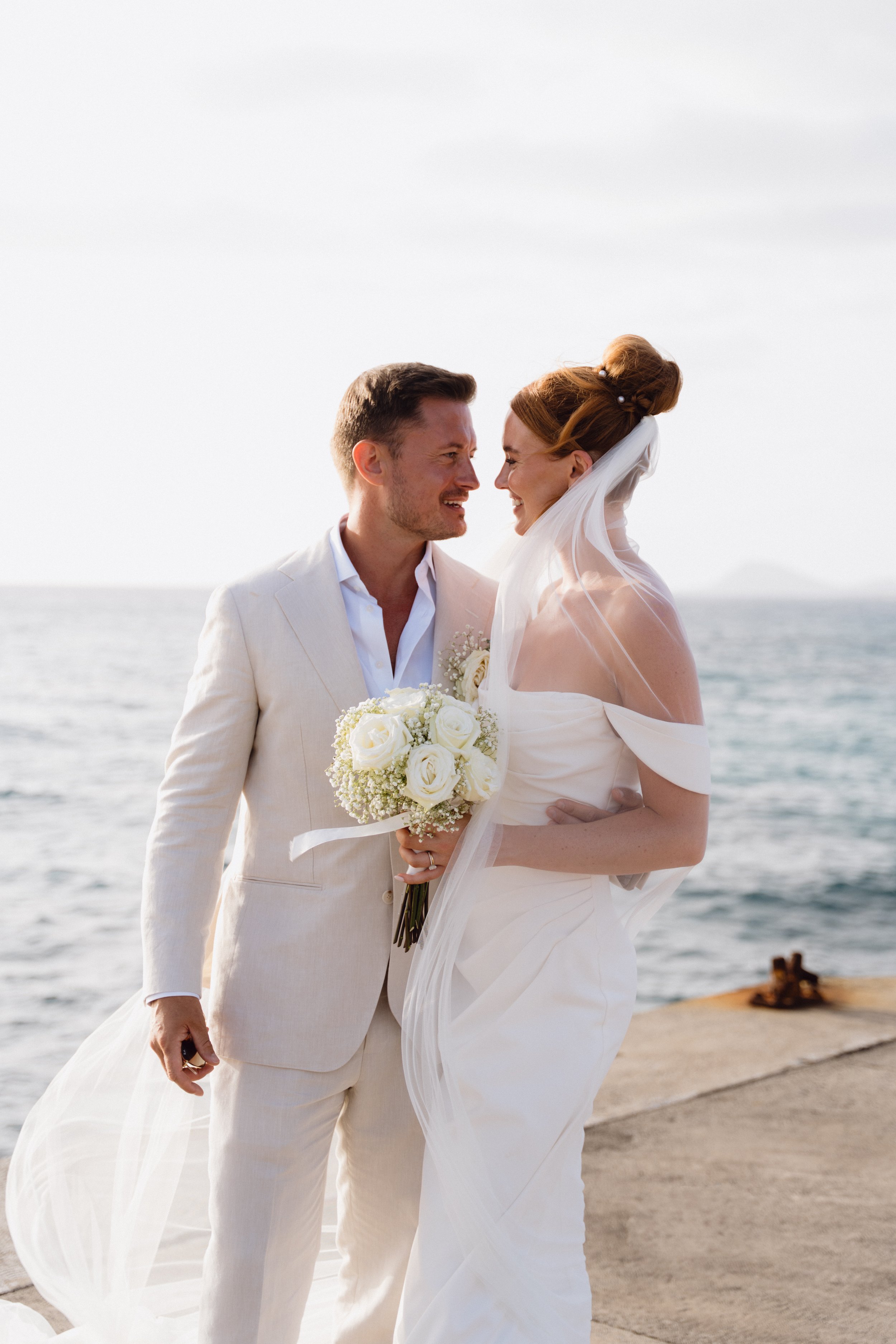 A bride and groom in wedding attire by the sea, sharing a happy moment, with the bride holding a bouquet of white roses.