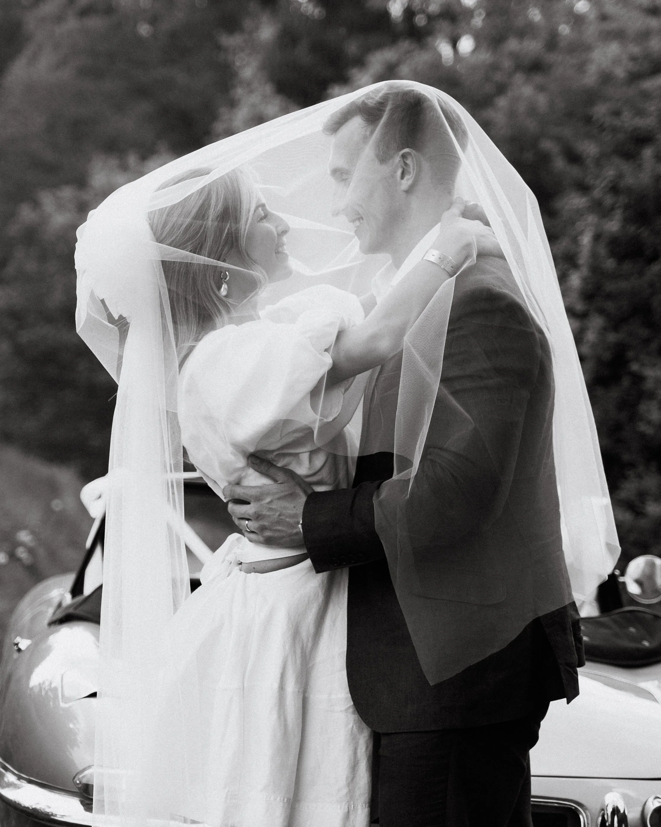 A couple on their wedding day under a veil, smiling and looking at each other in front of a car with a scenic background.