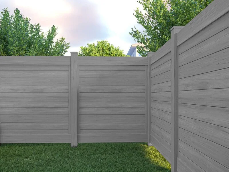 Gray wooden privacy fence beside green grass with trees and a house in the background under a partly cloudy sky.