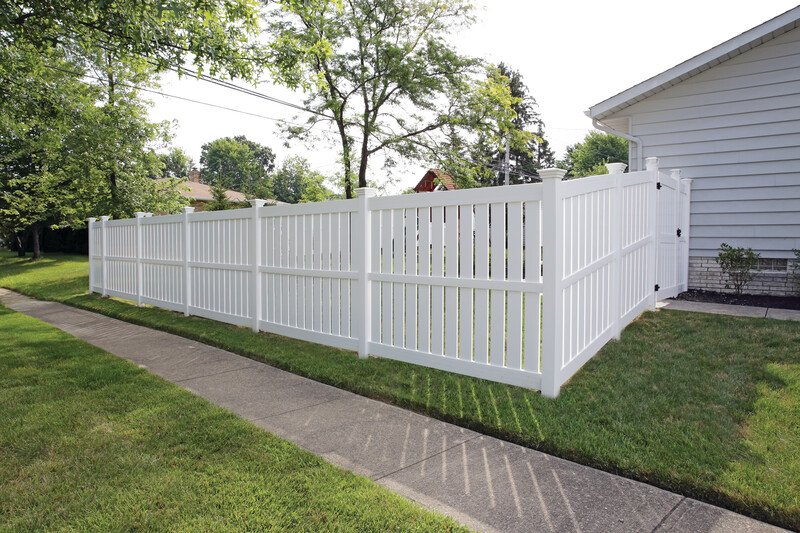 White privacy fence along a sidewalk with green lawn and trees in a residential yard