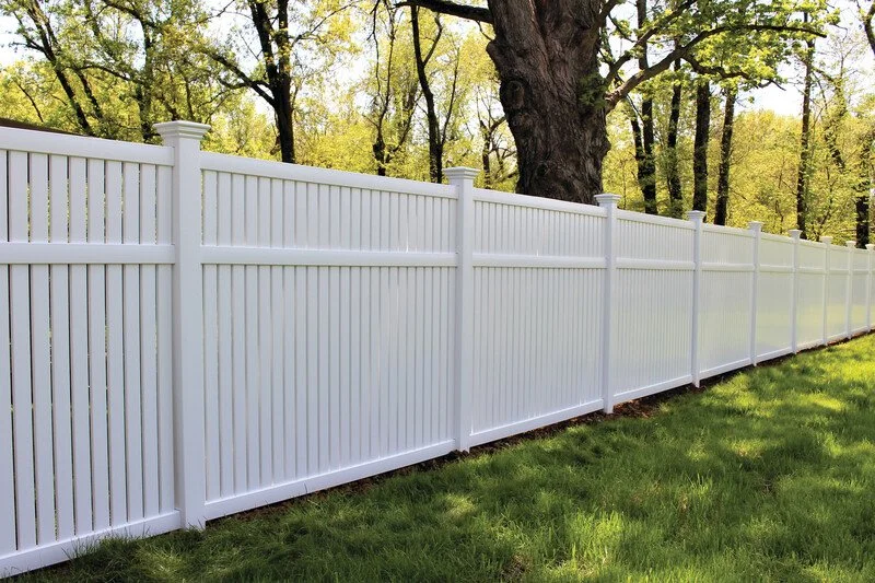 White privacy fence running along a grassy yard with trees in the background.