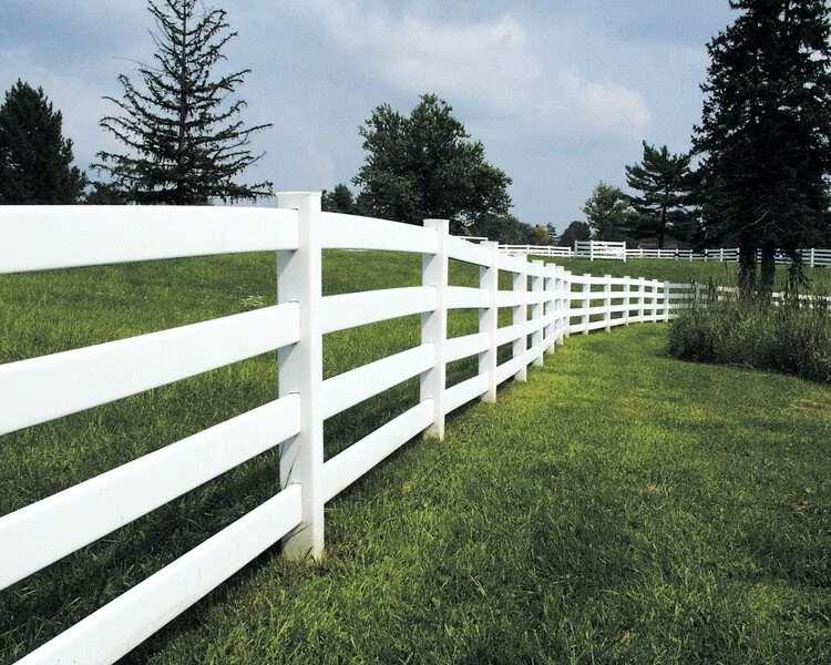 Green grassy yard with a white fence, forested hills, and snow-capped mountains in the background under a partly cloudy sky.