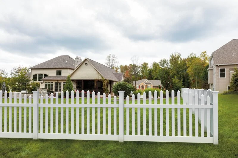 Residential backyard with white picket fence, green lawn, and two houses, one with a porch, under a cloudy sky