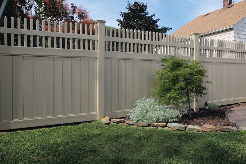 A beige privacy fence nearby a green lawn with a small shrub and flowering plant, in a backyard with a house in the background.