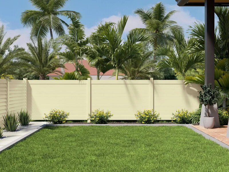 A backyard with a well-maintained green lawn, decorative plants, and yellow flowers along the fence. There is a white fence, and tropical palm trees are visible beyond the fence. A corner of a house with a potted plant is in the right foreground.