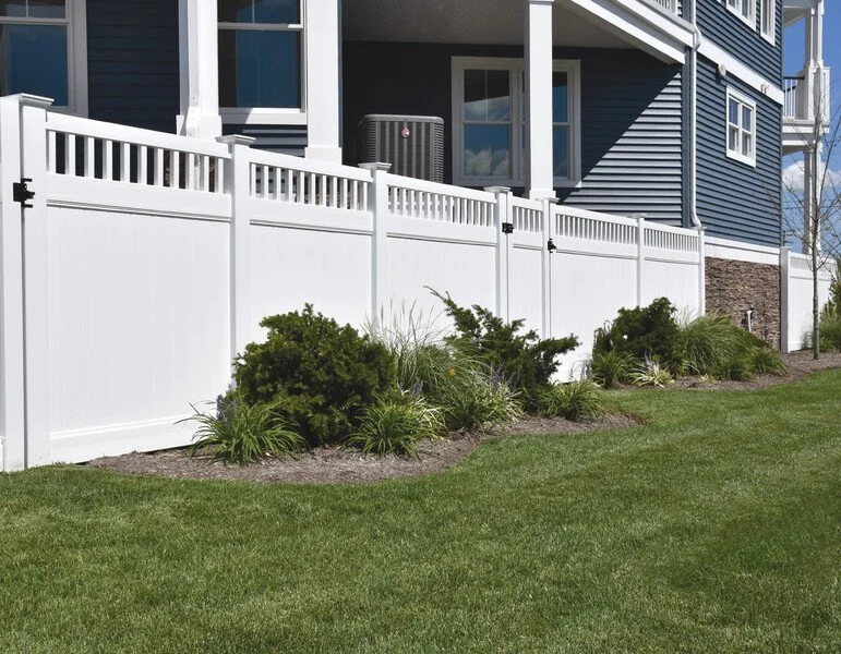 White privacy fence in front of a blue house with a landscaped yard including bushes and grass.