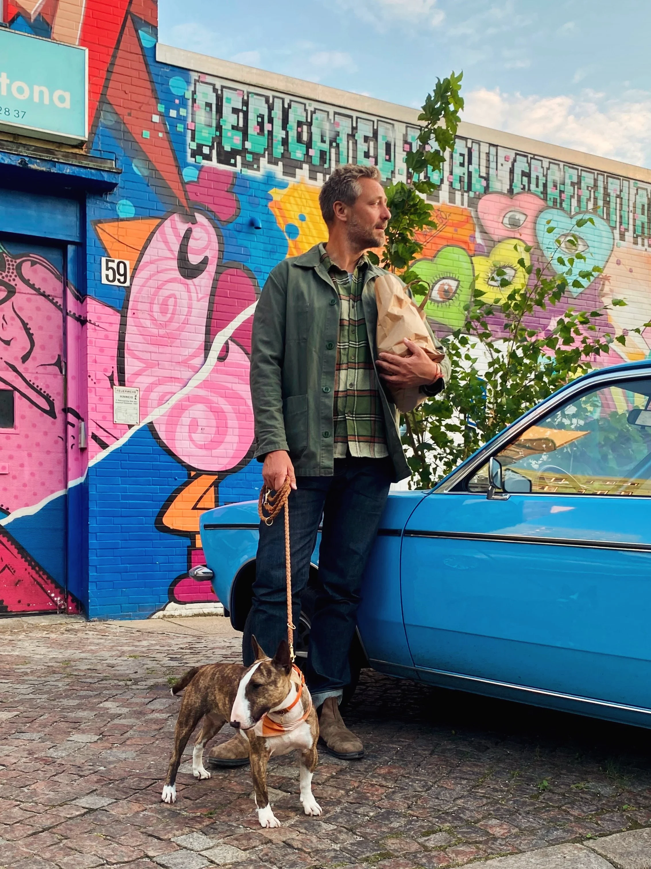 A man standing next to a blue vintage car, holding a leash attached to a brindle and white dog wearing an orange bandana. The man is in front of a colorful mural with graffiti art on a brick wall, featuring hearts, eyes, and vibrant patterns. The sky is partly cloudy.