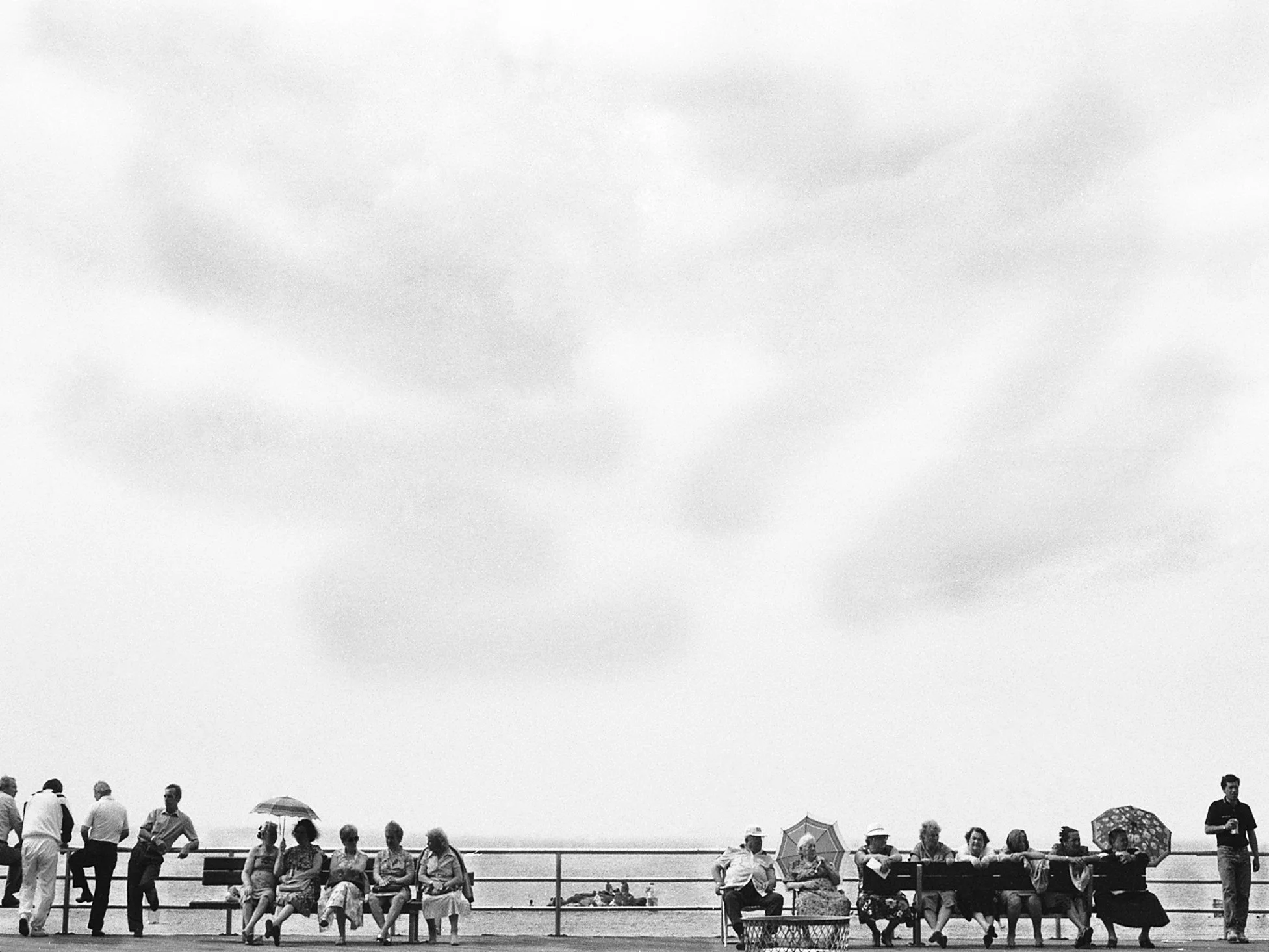 Black-and-white photograph of people seated along a seaside boardwalk beneath an open sky.