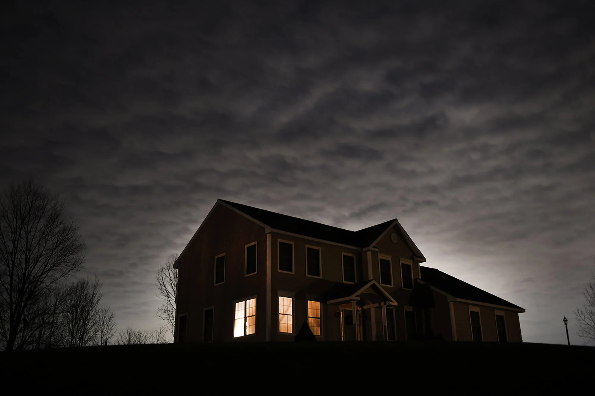 A silhouette of a large house at night with lit windows against a cloudy sky.