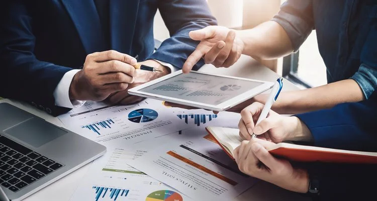 Three Business People Planning Strategically with a notebook and pen, an ipad, a computer, and papers on the table.