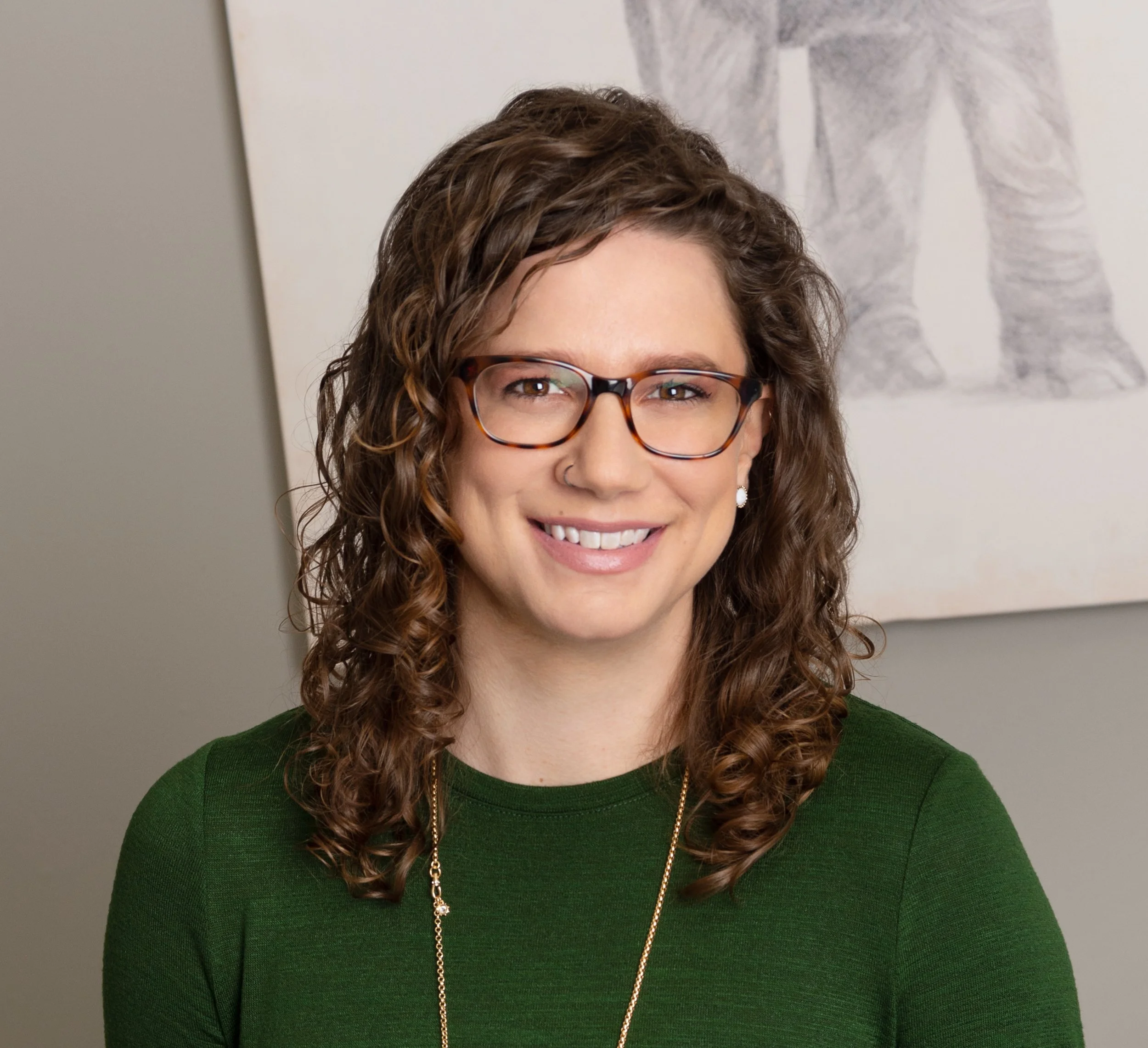 A woman with curly brown hair, wearing glasses and a green top, smiling in front of a white abstract artwork.