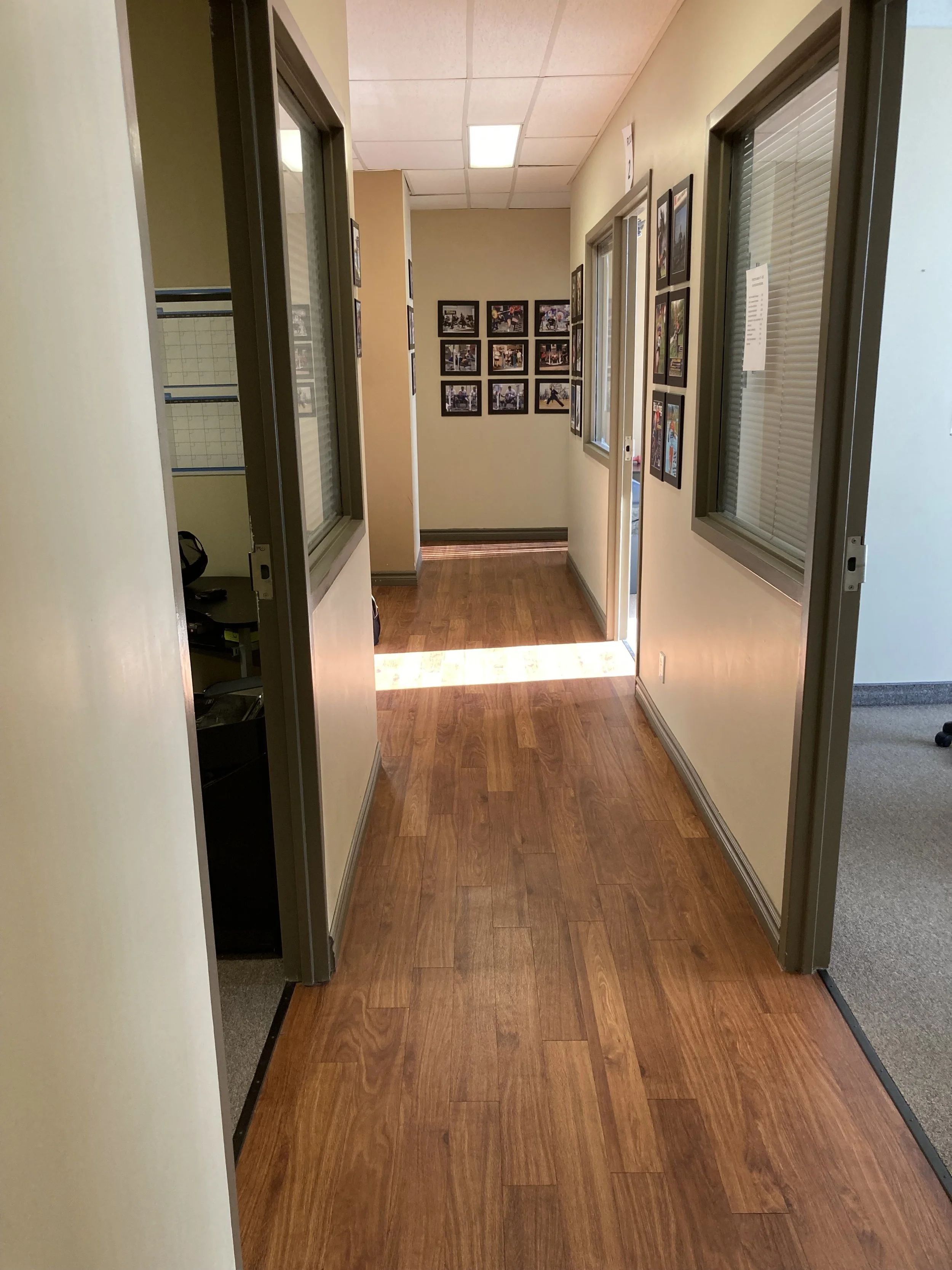 A hallway with windows on the right and framed pictures on the walls, with a hardwood floor and ceiling panels.