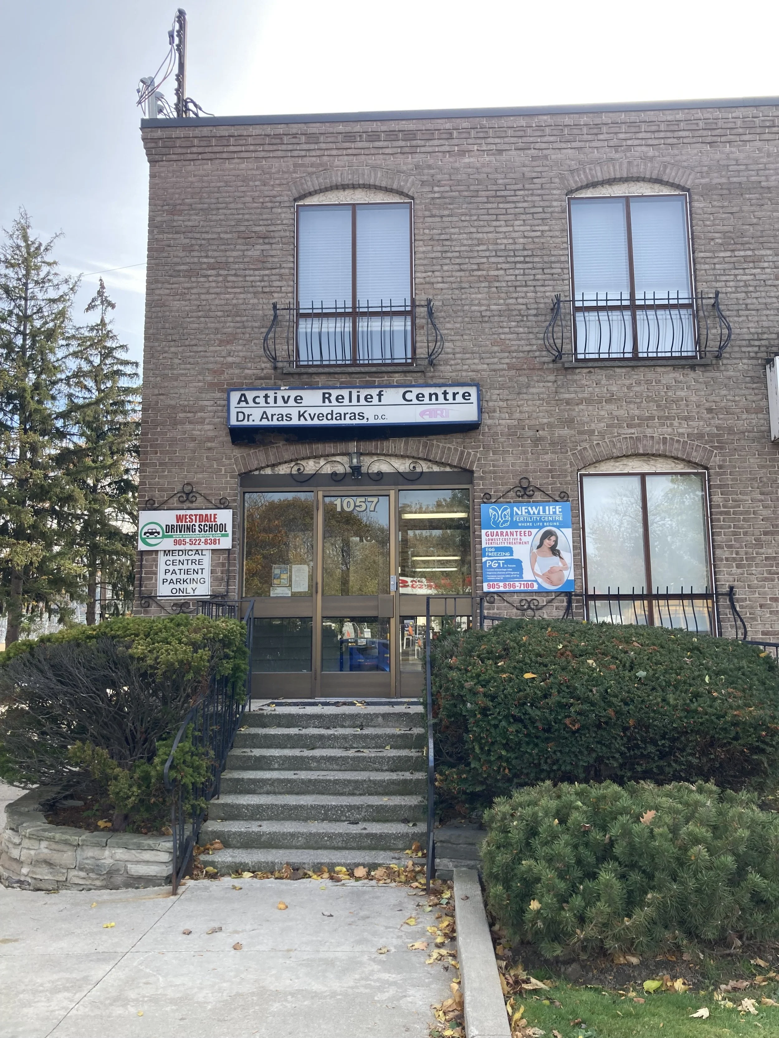 Brick building with sign for Active Relief Centre and Dr. Aras Kvedaras, DC. Entrance with stairs, surrounded by bushes and trees, and multiple windows.