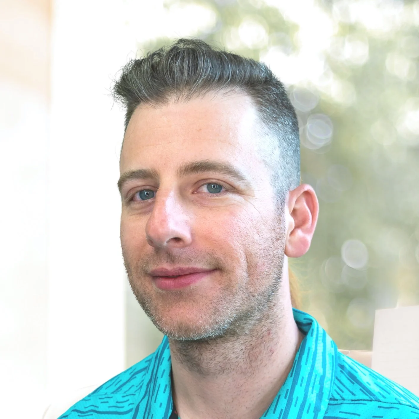 A handsome man with blue eyes, short dark hair, and a light beard, wearing a teal shirt with a subtle pattern, smiling slightly, with a blurred outdoor background and soft lighting.