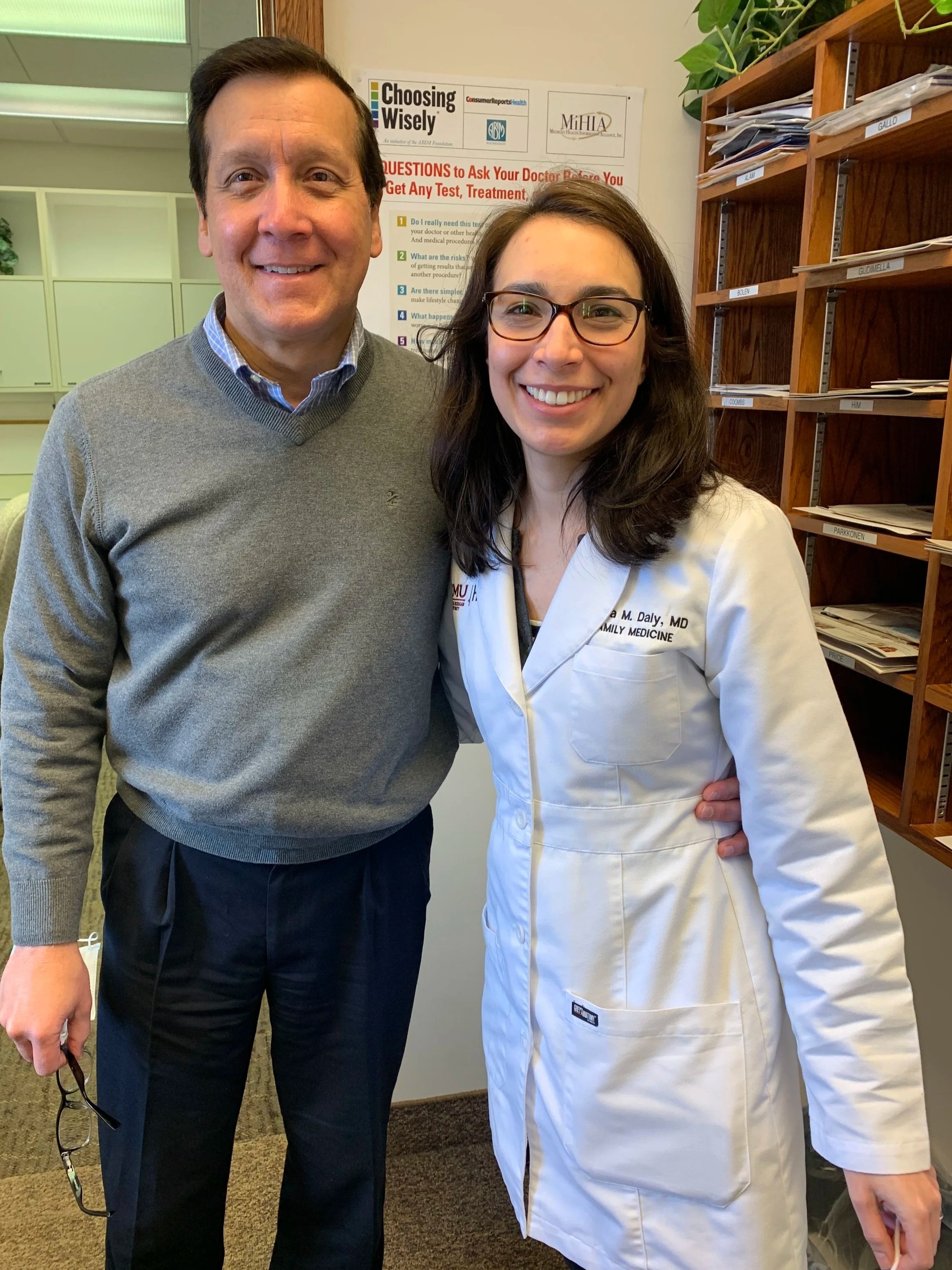 A man and woman smiling and standing close together in an indoor office setting. The man is wearing a gray sweater over a blue collared shirt and black pants, holding glasses in his left hand. The woman is wearing glasses and a white medical coat with a name badge, and has her arm around the man's waist. Behind them are shelves with folders and a poster about health questions.