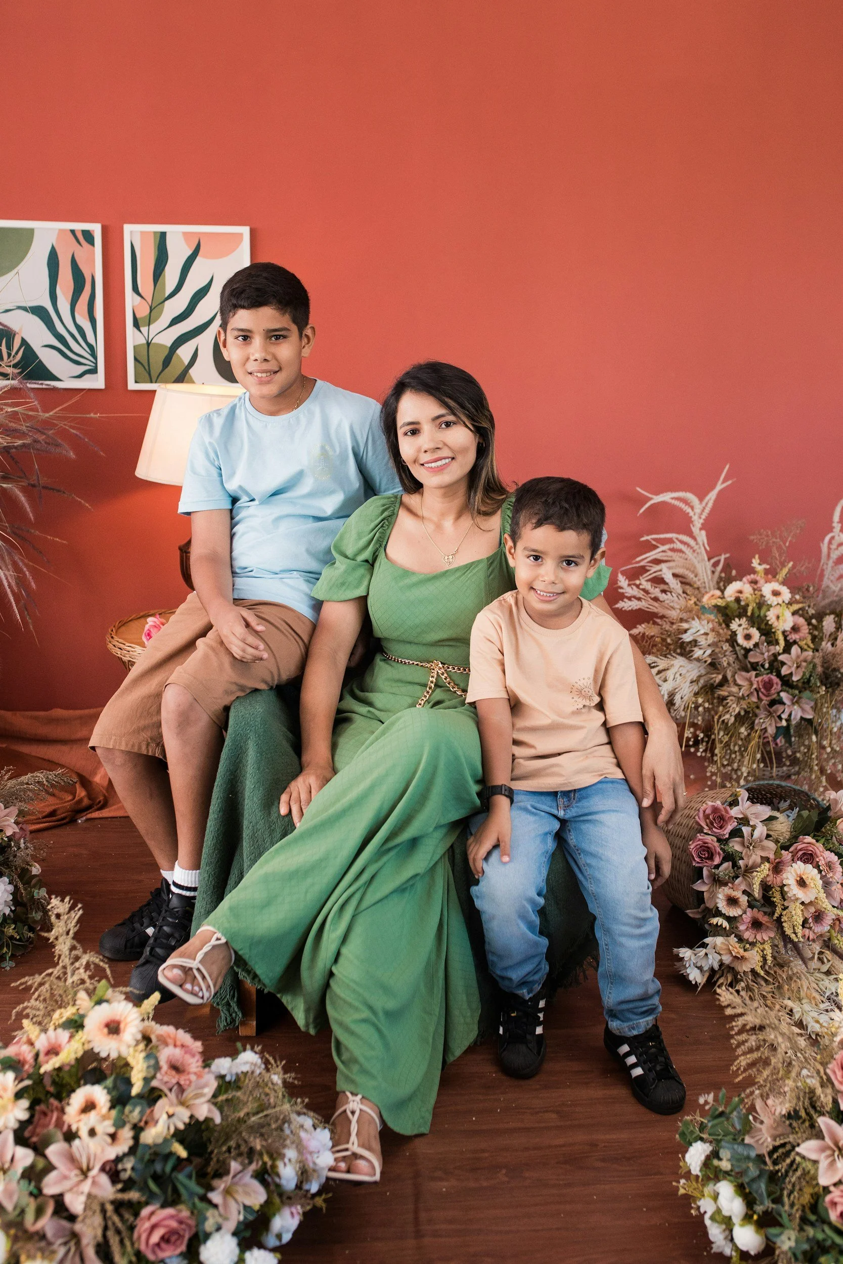 A woman sitting on a chair with two young boys on her lap, surrounded by floral arrangements, against a red wall with framed botanical artwork.
