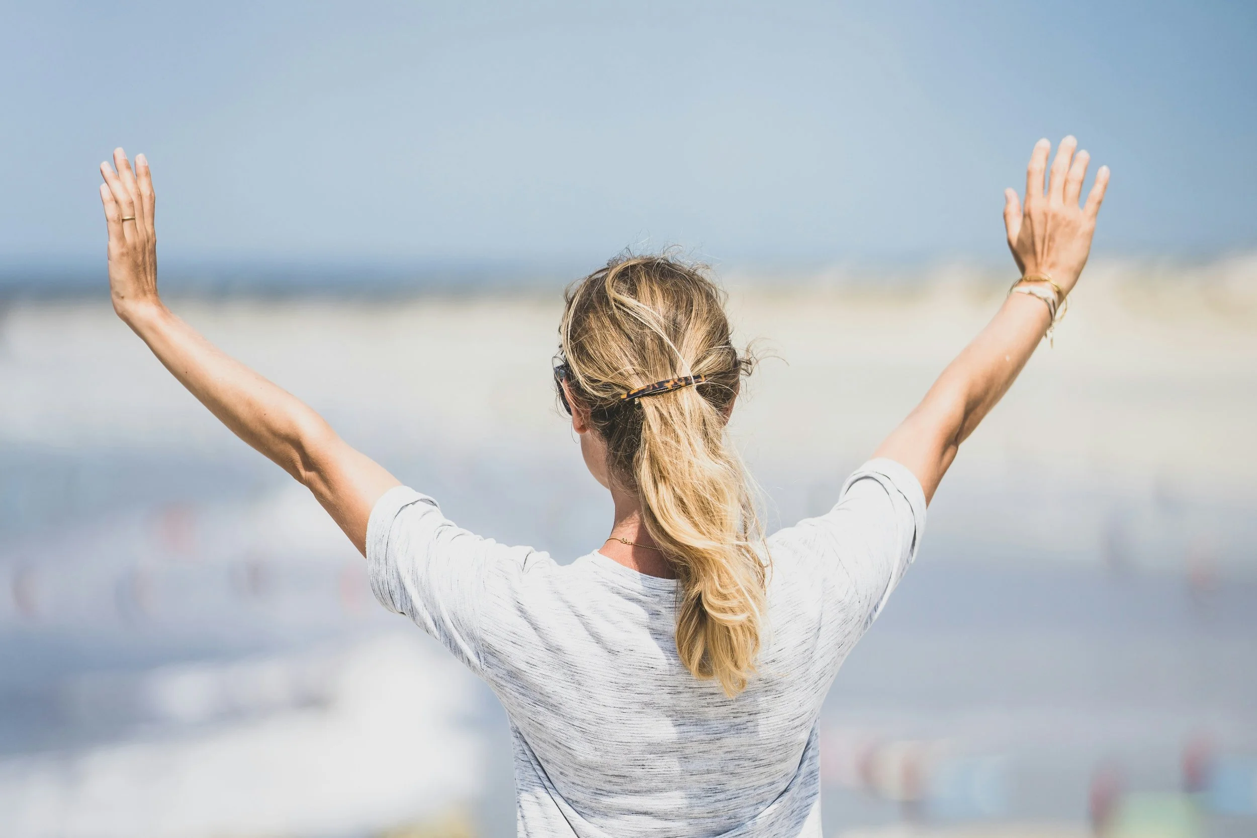 A woman with blonde hair tied back, wearing a light gray T-shirt, standing on the beach with her arms raised, facing the ocean.