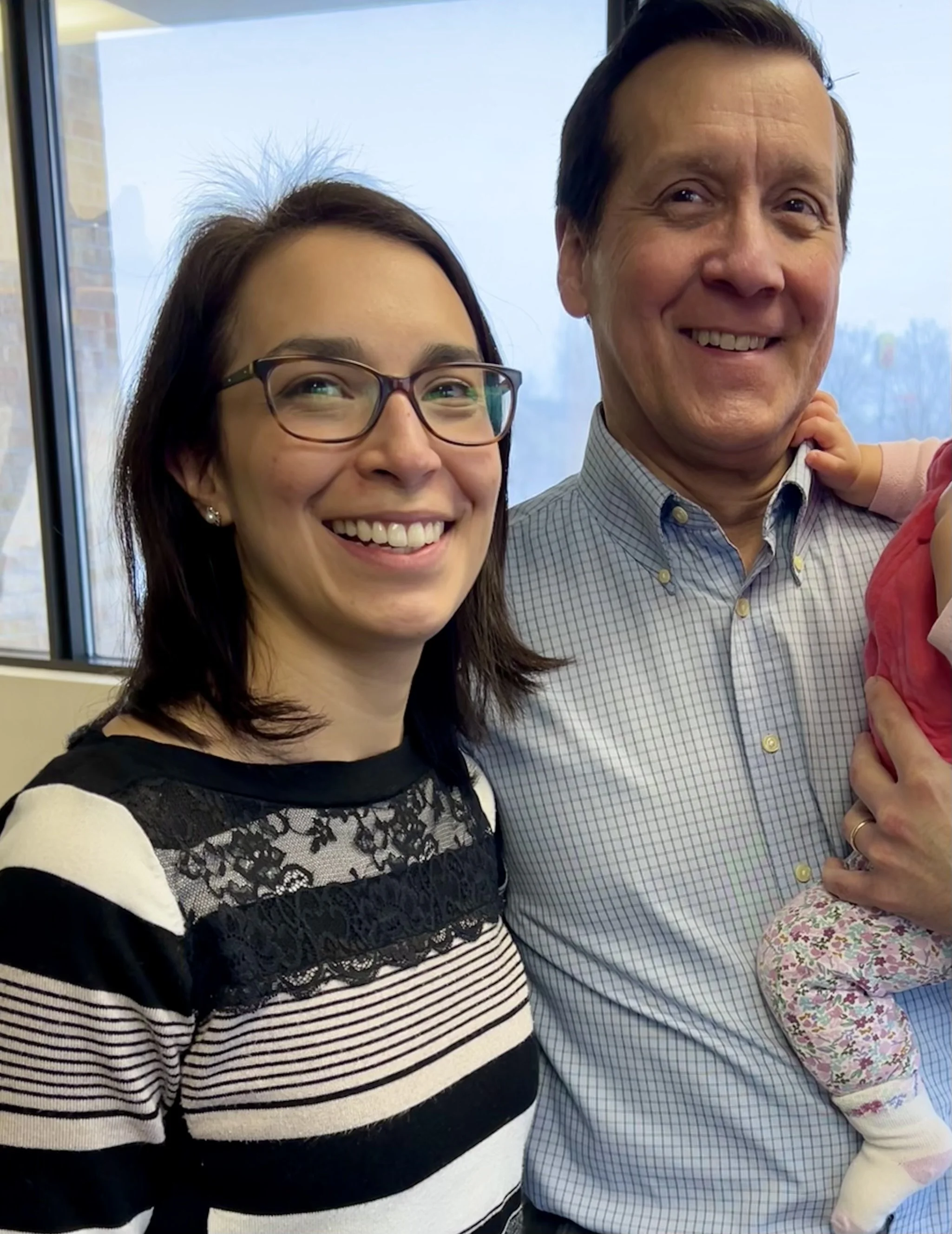 Smiling woman with glasses and a man holding a child in a bright room with a window in the background.