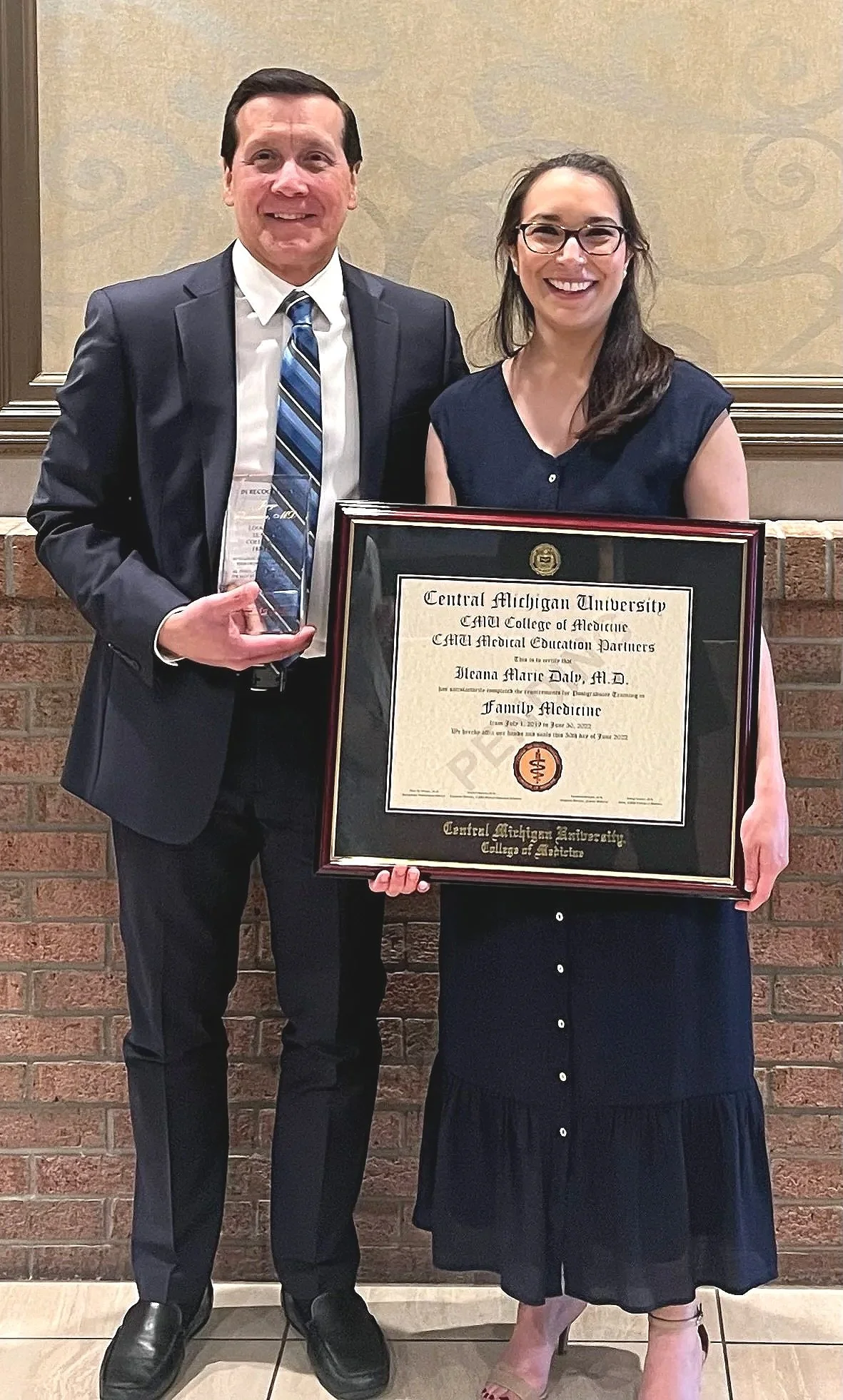 A man and a woman dressed in formal attire, standing in front of a brick wall. The woman is holding a framed diploma from Central Michigan University College of Medicine, and the man is holding a clear award made of glass or crystal.