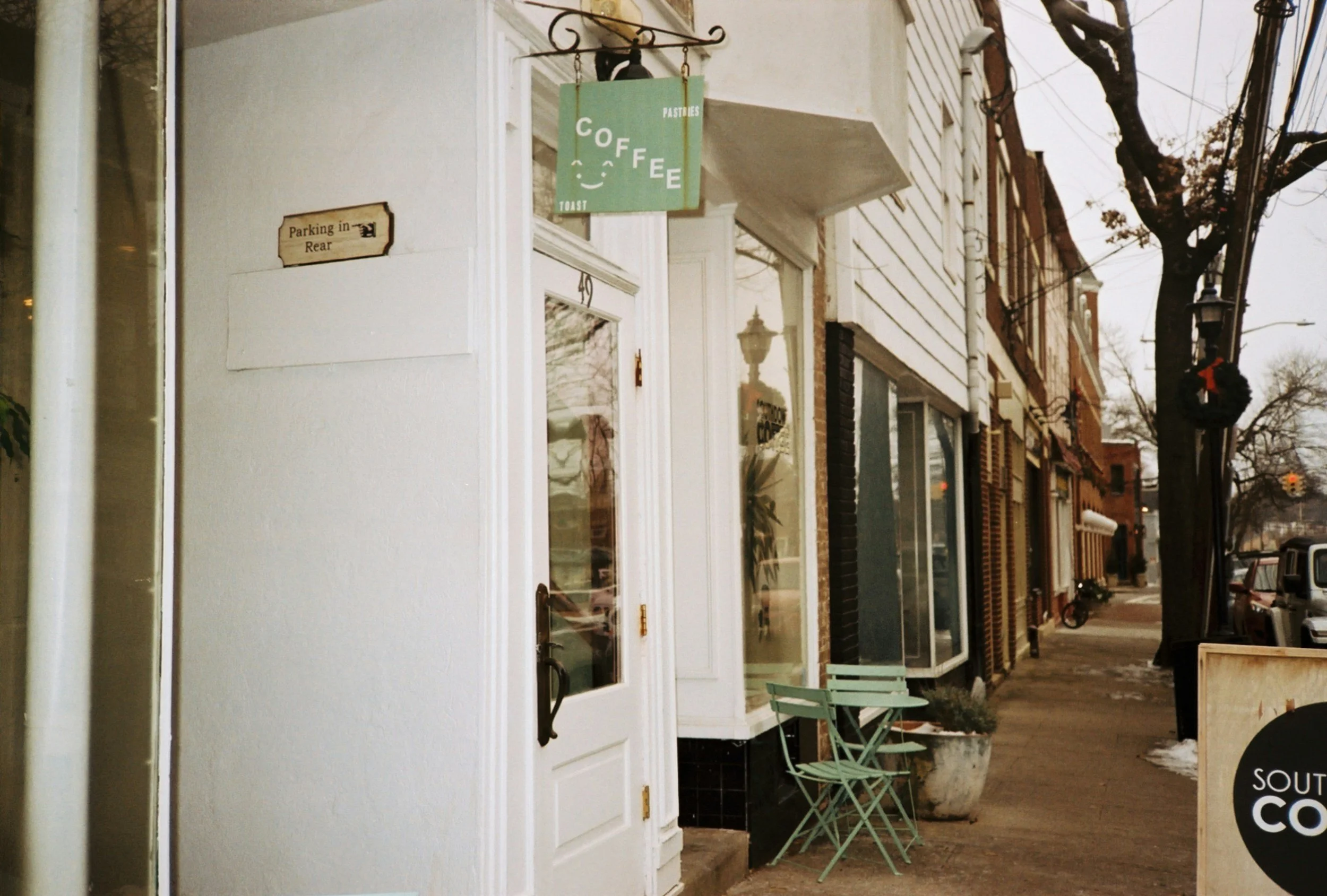 Street view of small coffee shop with a green sign that says 'Coffee' and a smiling face, outdoor seating with green chairs, and a sidewalk with a few patches of snow.