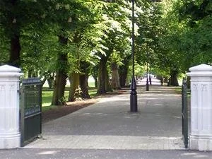 A park pathway lined with green trees, with black lampposts and white stone pillars at the entrance.