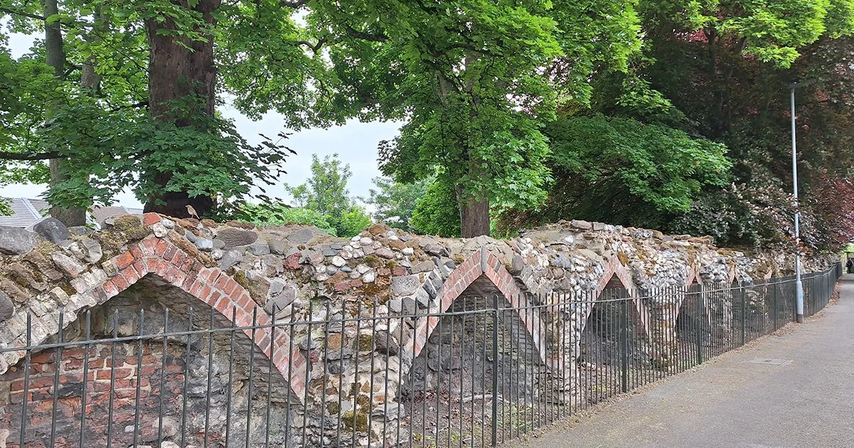 King's Lynn's old town walls.  
A historic stone and brick wall with a zigzag pattern, topped with greenery from trees, and a black metal fence along a sidewalk.
