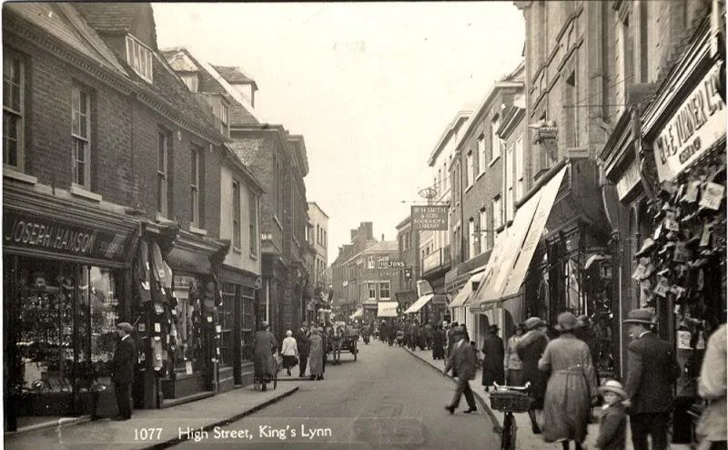 Historical black and white photo of High Street in King's Lynn, showing pedestrians, shops, and horse-drawn carriages.