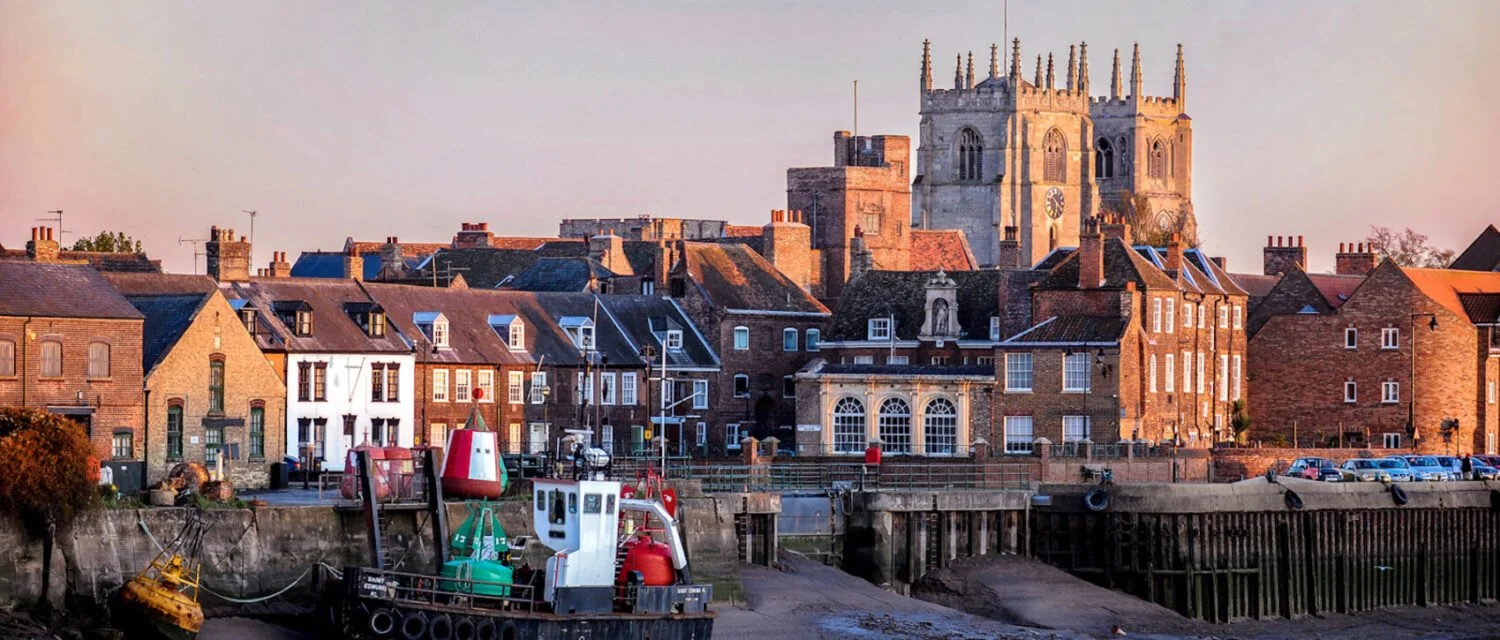 View of a coastal town with a historic cathedral in the background, boats docked at a harbor, and traditional brick buildings with chimneys in the foreground during sunset.