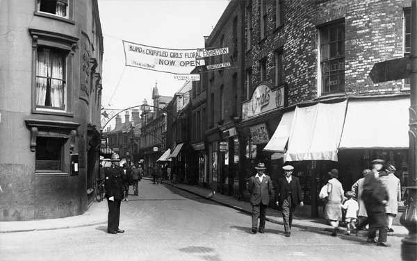 Black and white photo of a city street with pedestrians walking on the sidewalk, shops with awnings, and a banner hanging across the street advertising a floral exhibition.