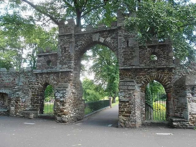 Old stone gate in a park, with archways and a pathway leading through it, surrounded by green trees.