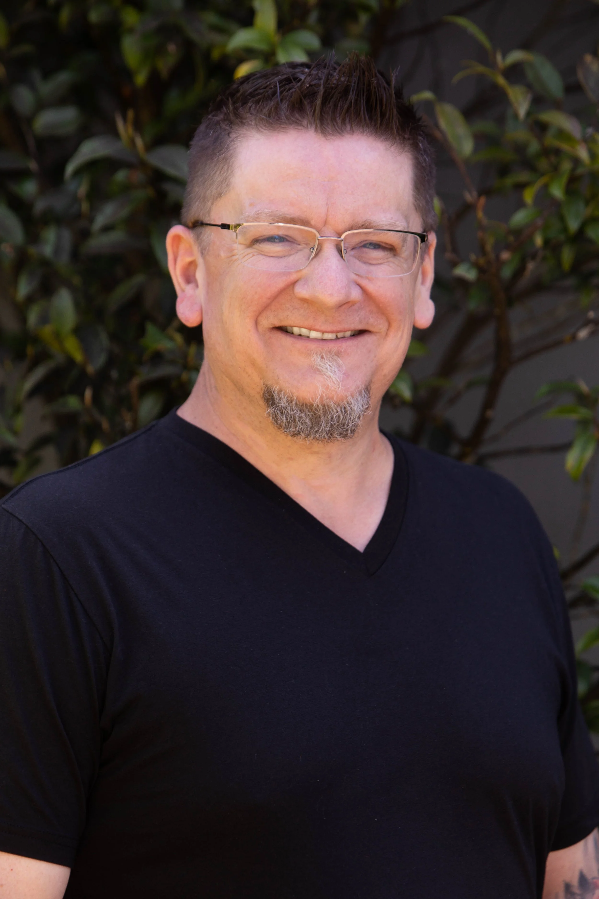 Smiling person wearing a black cap and a black shirt with a goatee, standing outdoors in front of a gray wall.
