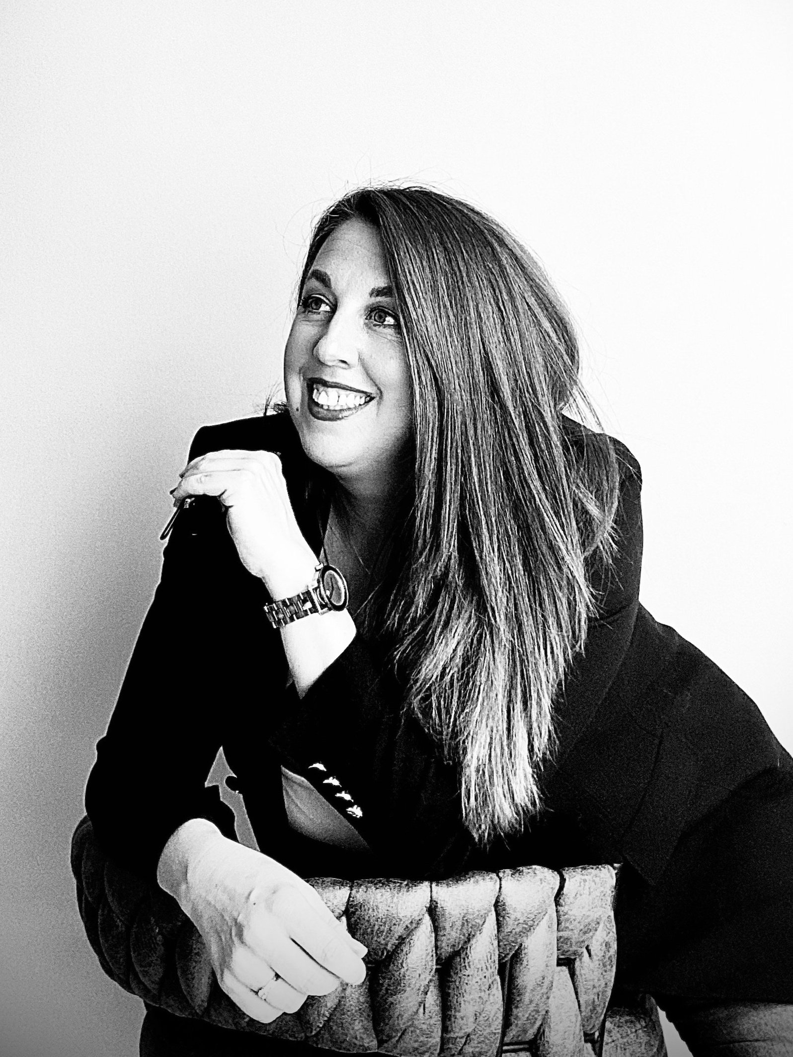 Black and white photo of a woman with long hair smiling, leaning on a textured chair, wearing a watch and a dark blazer.