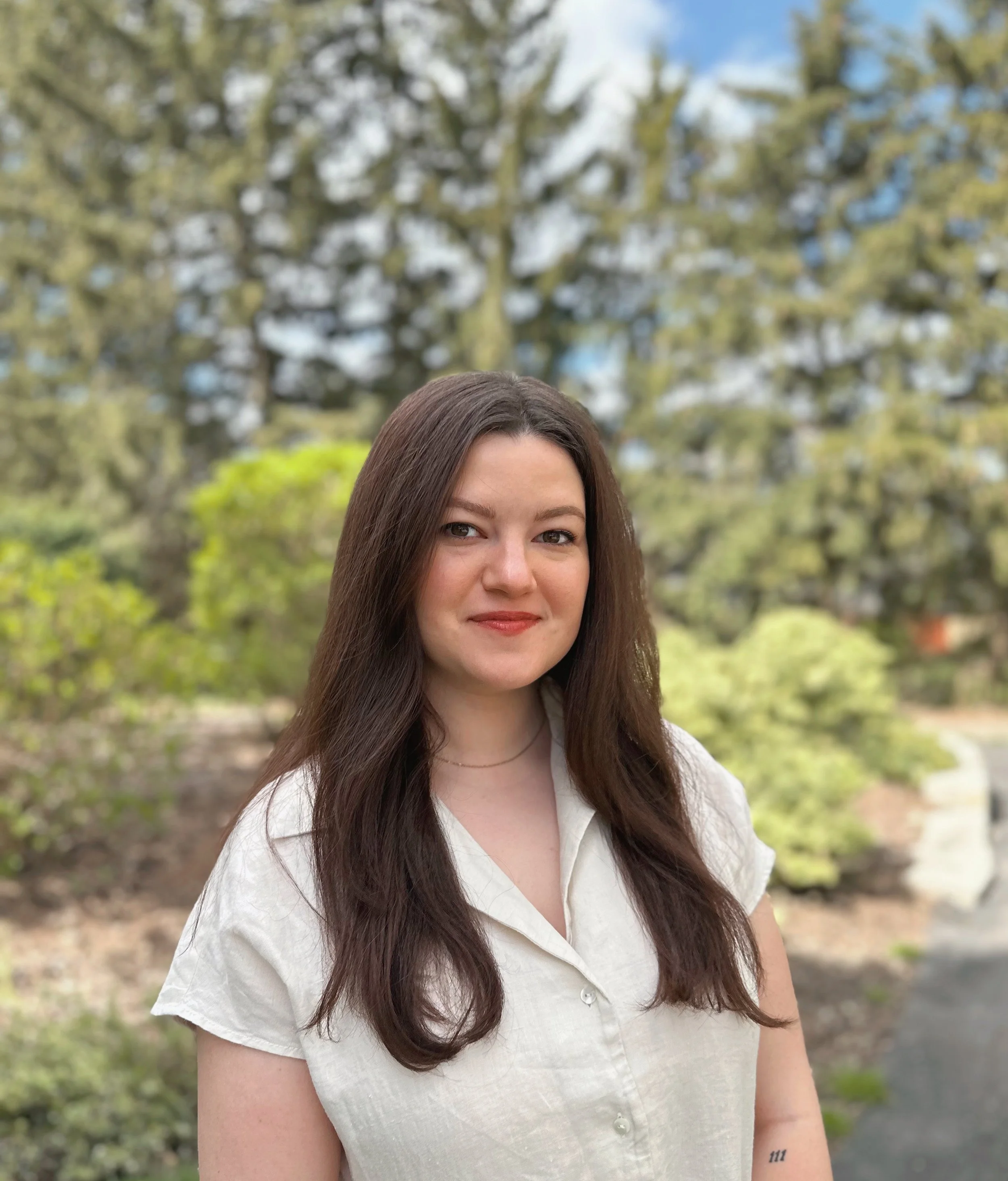 A woman with long brown hair wearing a white shirt standing outdoors against a background of trees and sky