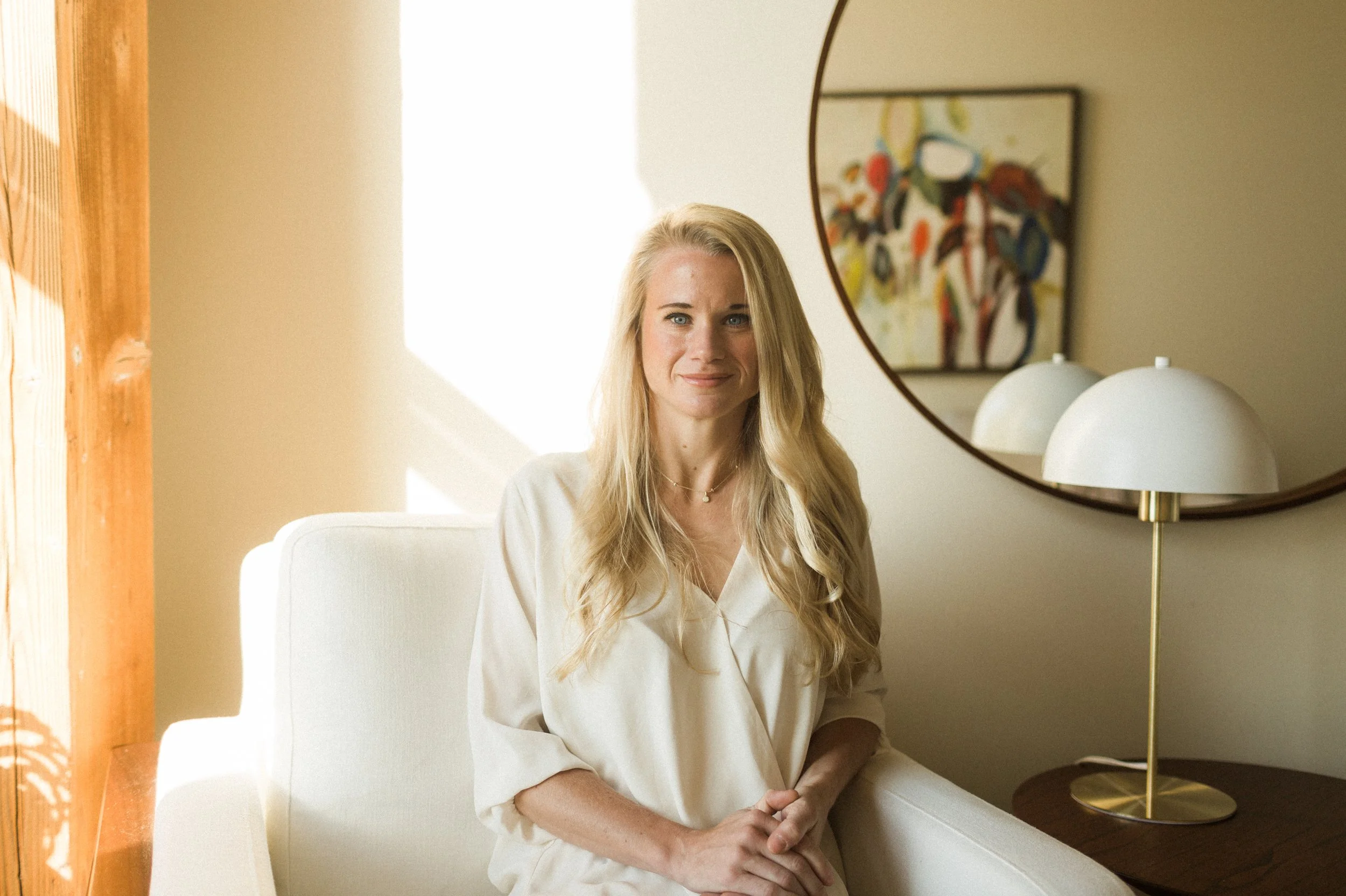 A woman with long blonde hair sitting on a white sofa in a brightly lit room, with a colorful painting on the wall behind her and a globe-shaped lamp on a wooden side table.