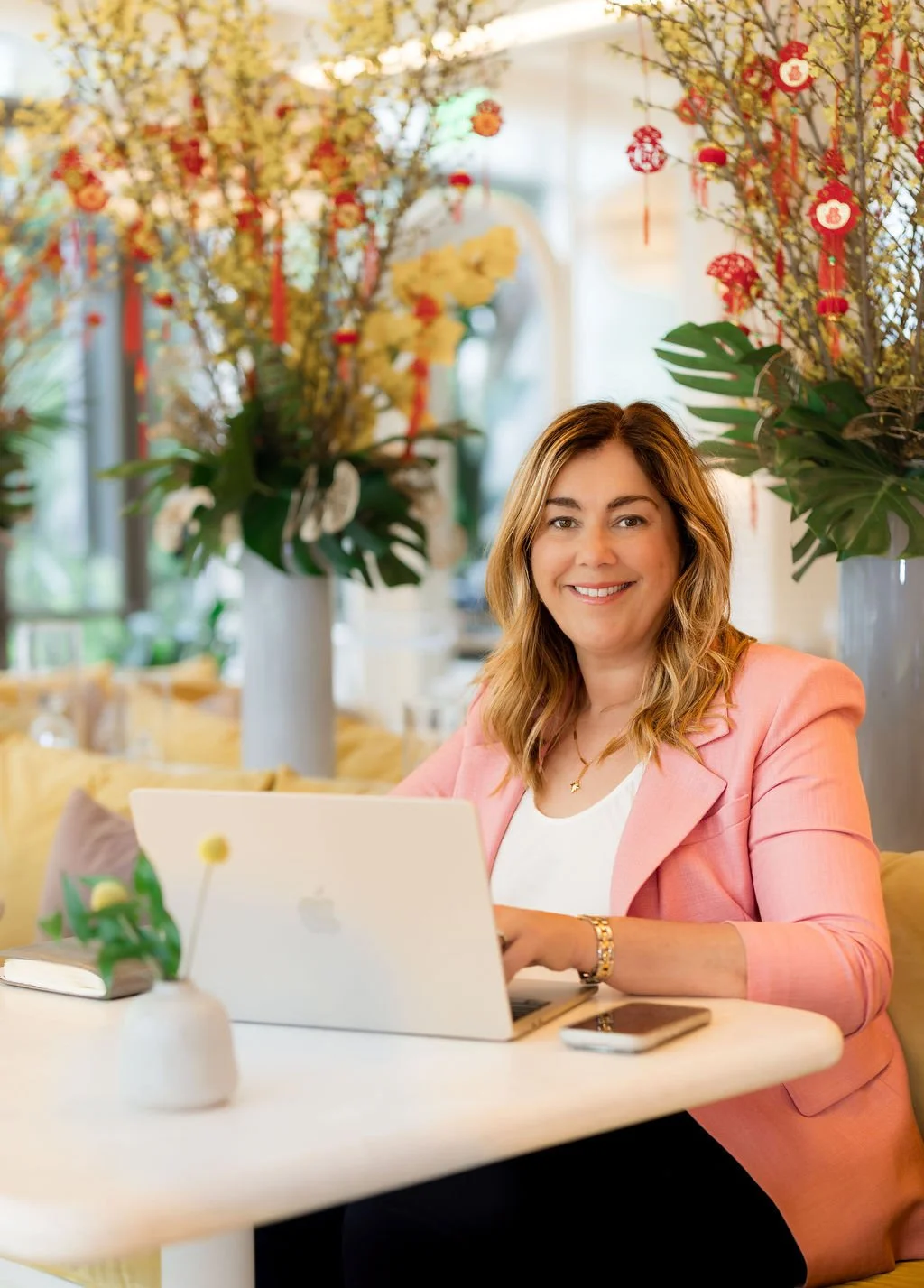 A woman sitting at a table with a laptop and a smartphone, smiling, in a brightly decorated room with large floral arrangements and colorful hanging decorations.