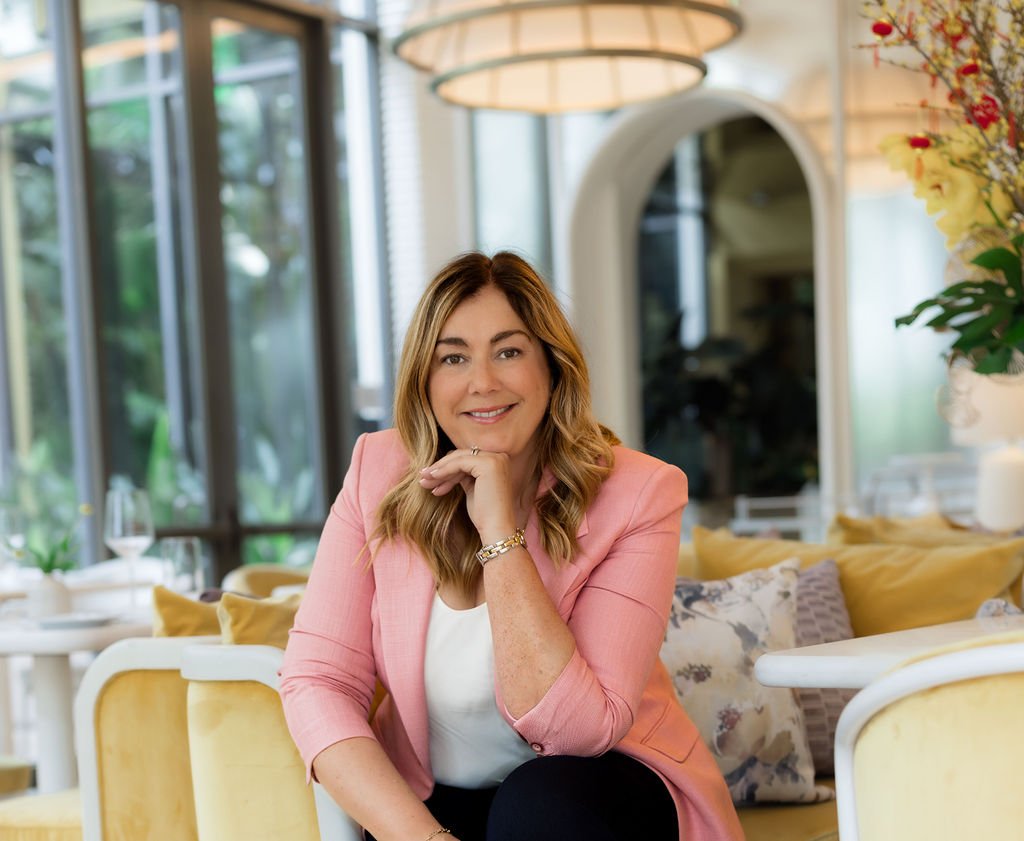 A woman with shoulder-length blonde hair in a pink blazer sitting in a brightly lit restaurant or cafe with yellow cushioned seating and large windows showing greenery outside.