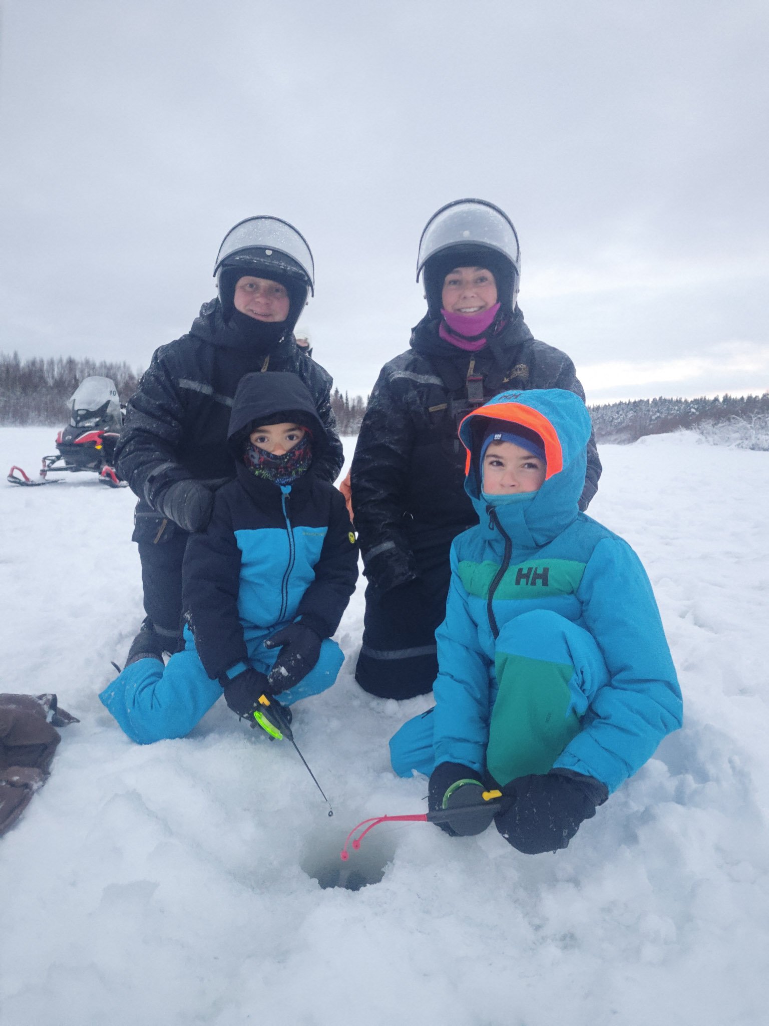 Four people in winter clothing and helmets kneeling and sitting in snow with a snowmobile in the background, outdoors with a snowy landscape and cloudy sky.
