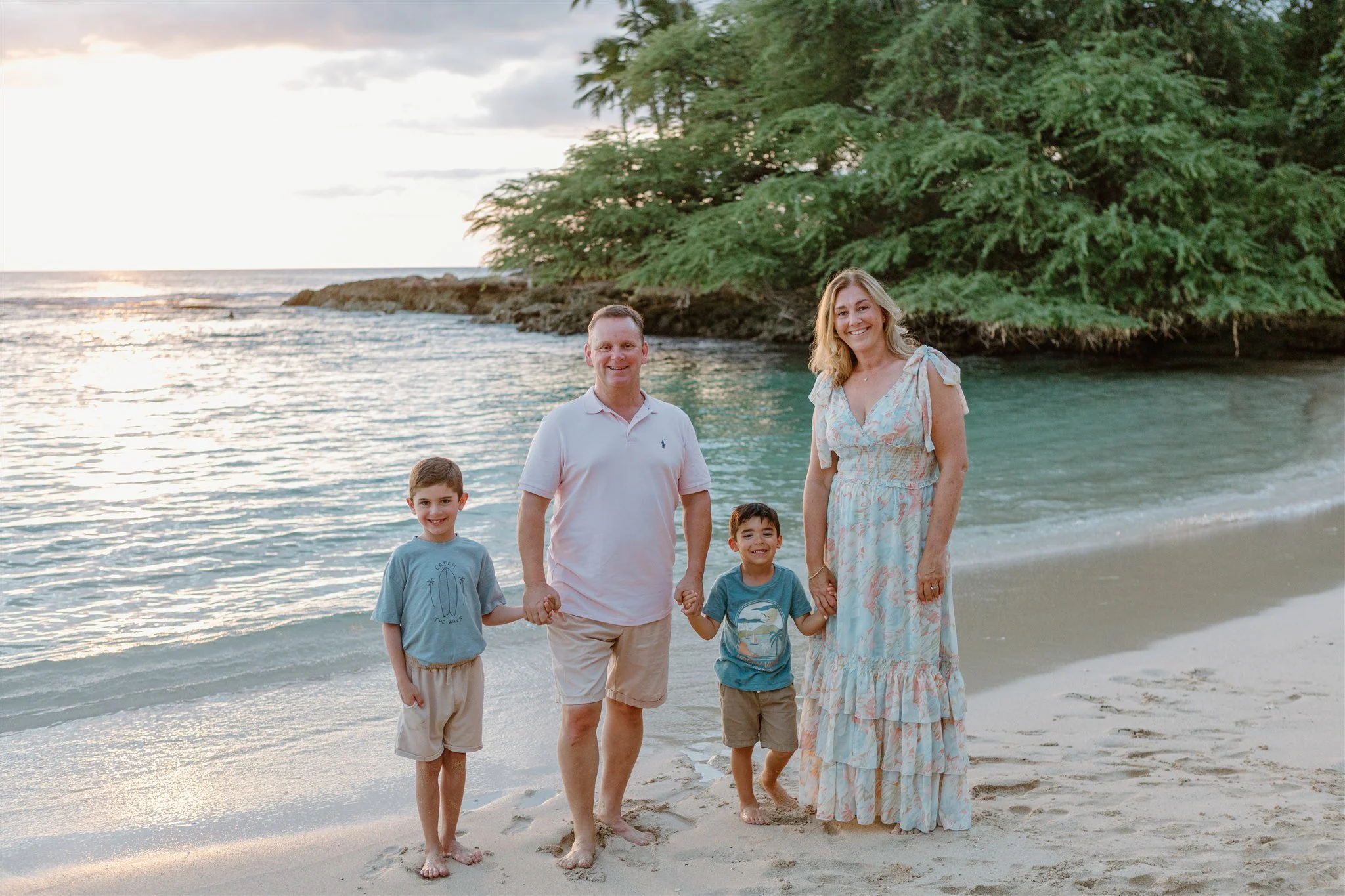 A family of five stands on the beach holding hands, with the ocean and a large green tree in the background during sunset. The family includes two boys, a man, a woman, and a young girl.