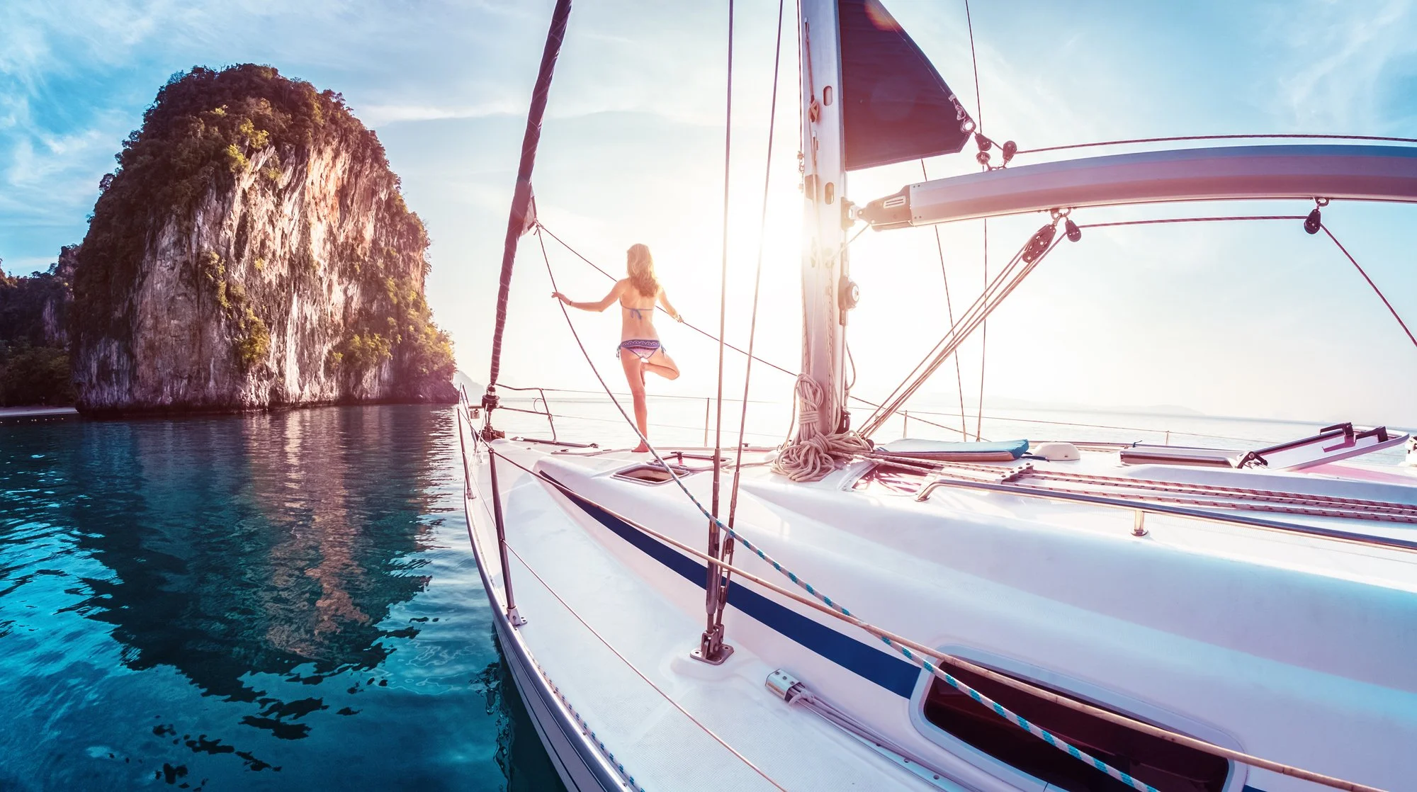 A woman in a bikini standing on a sailboat, holding the rigging, with a large rocky island in the background and clear blue water surrounding the boat.