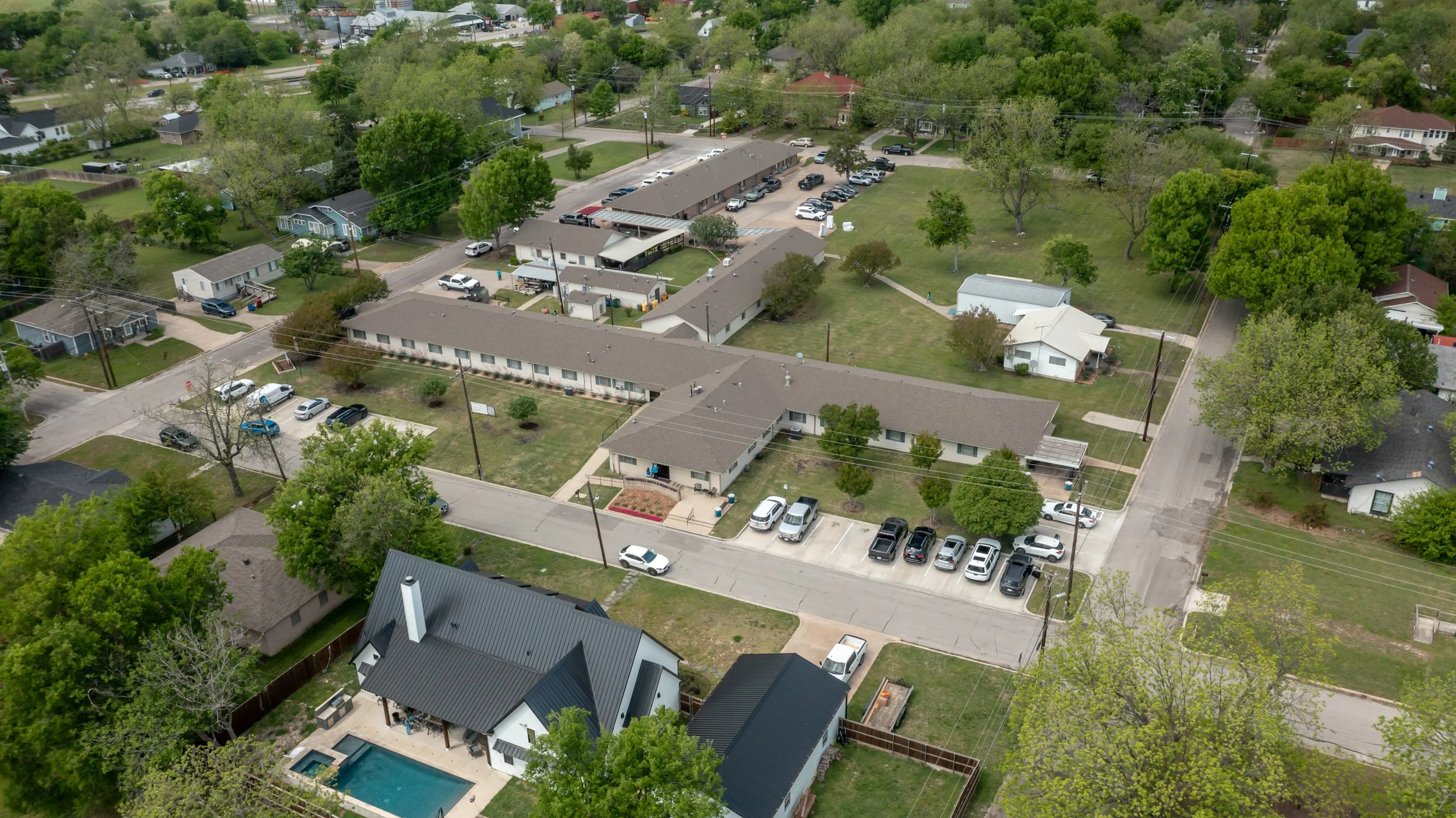 Aerial view of a suburban neighborhood with residential houses, parking lots, green trees, and a large courtyard complex with multiple buildings and a yard.