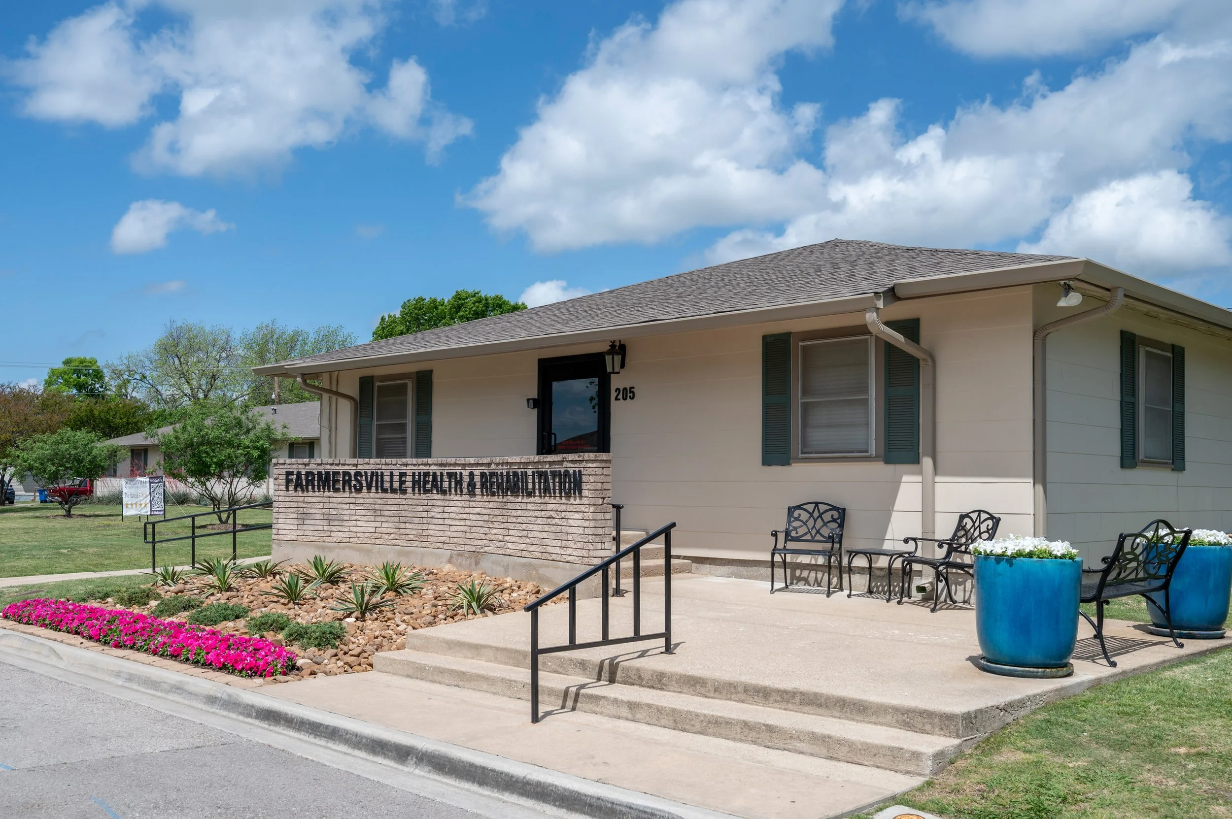 Exterior view of Farmer'sville Health & Rehabilitation building with a sign, benches, and planters, under a partly cloudy sky.