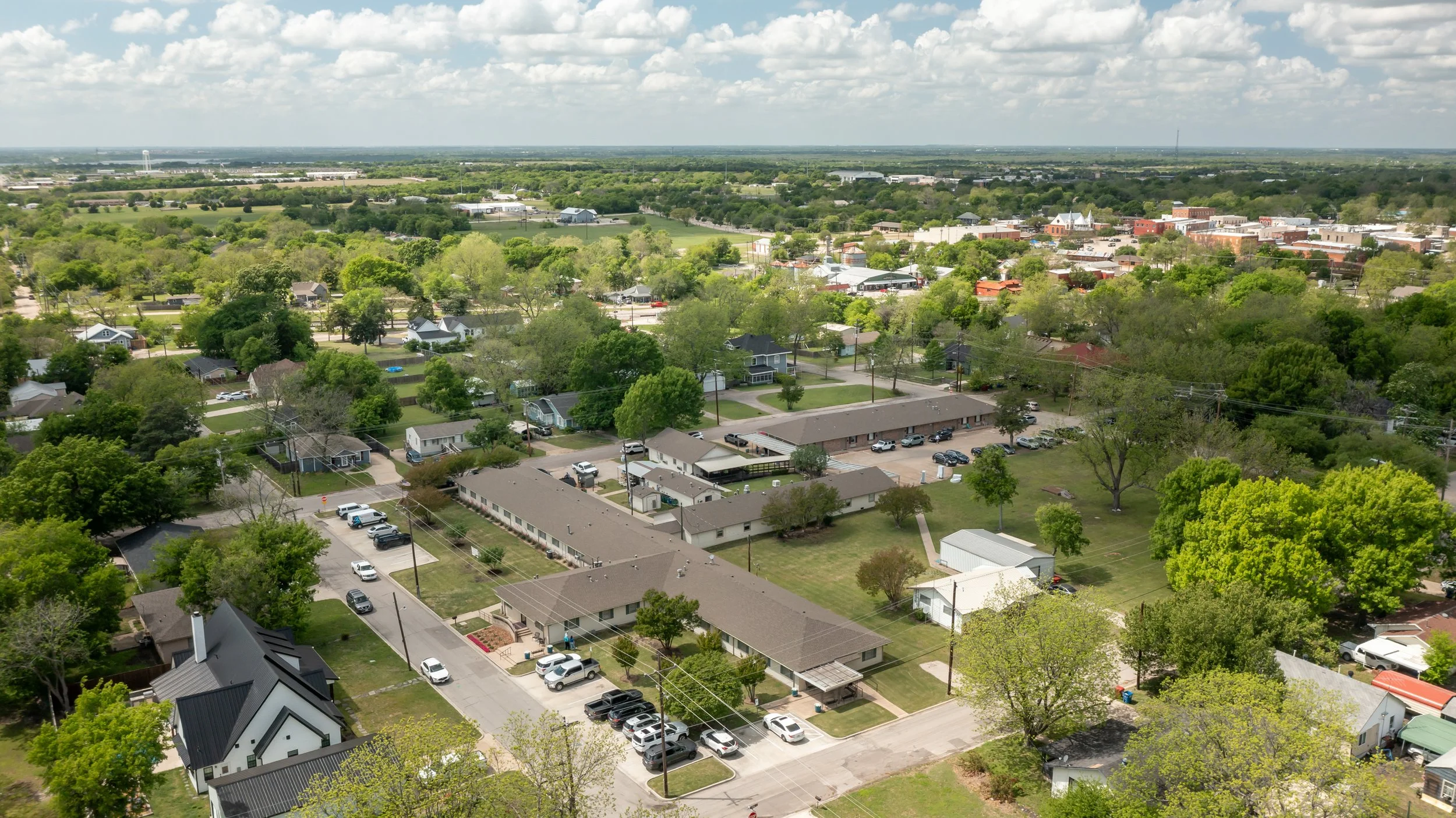 Aerial view of a suburban neighborhood with houses, trees, and a large building with a parking lot, under a partly cloudy sky.
