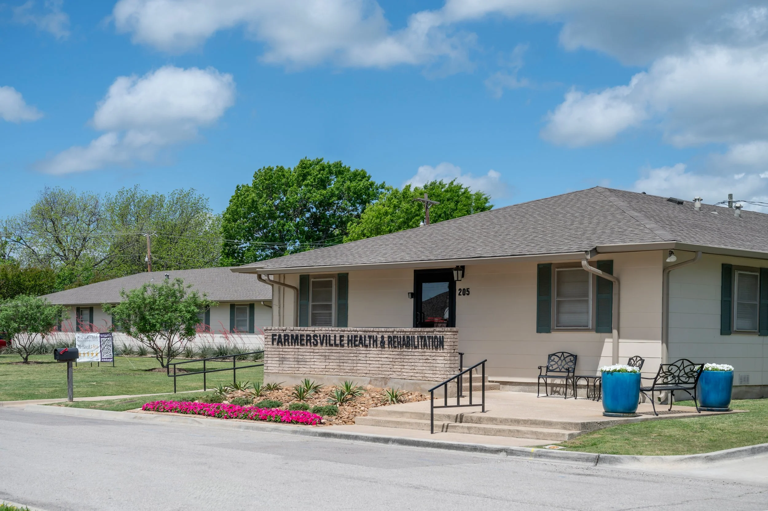 Exterior of Farmer'sville Health & Rehabilitation facility with benches and planters, surrounded by greenery under a blue sky