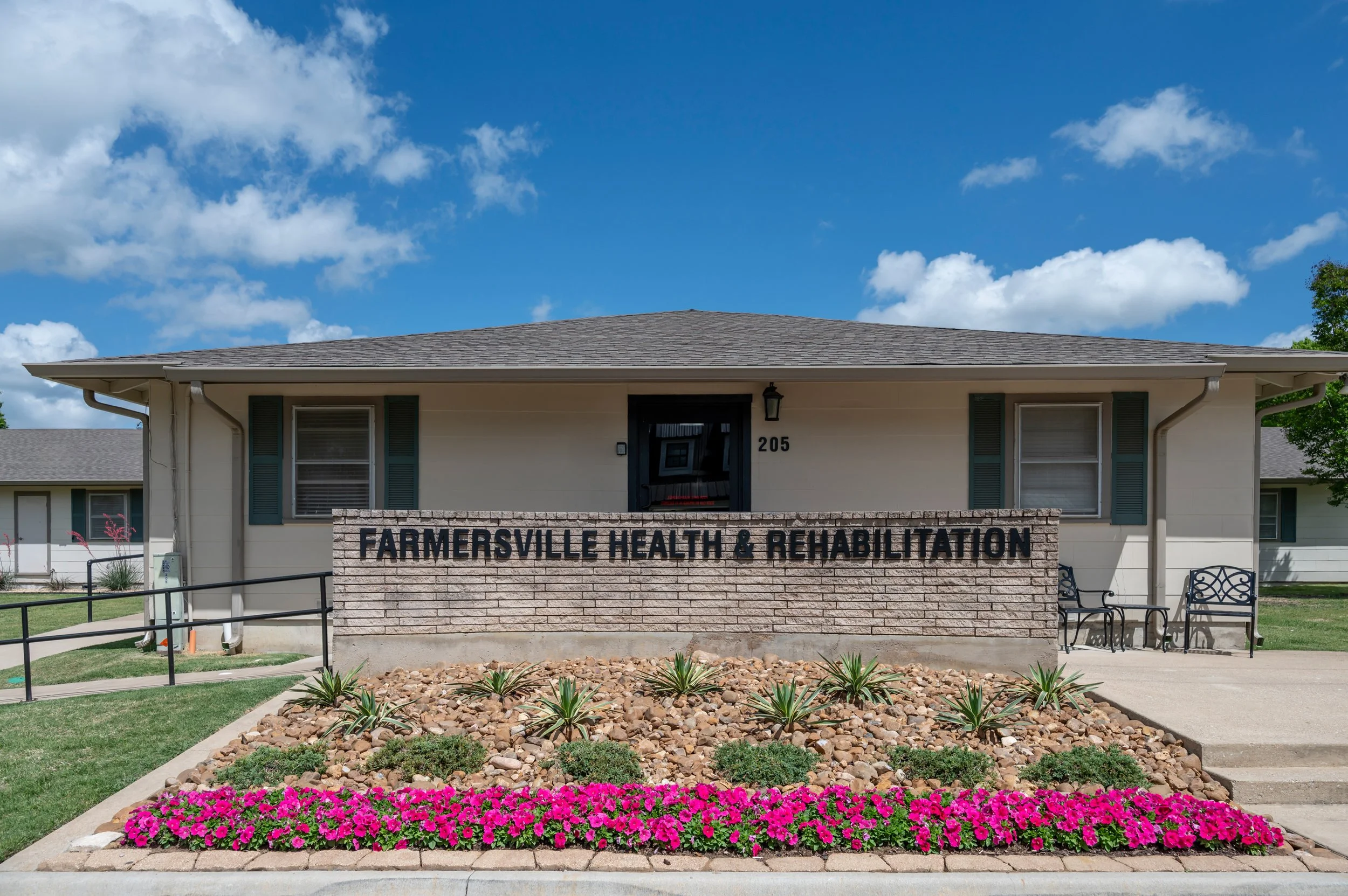Exterior view of Farmersville Health & Rehabilitation facility with a brick sign and flower bed in front, and a building with windows and a gray roof.