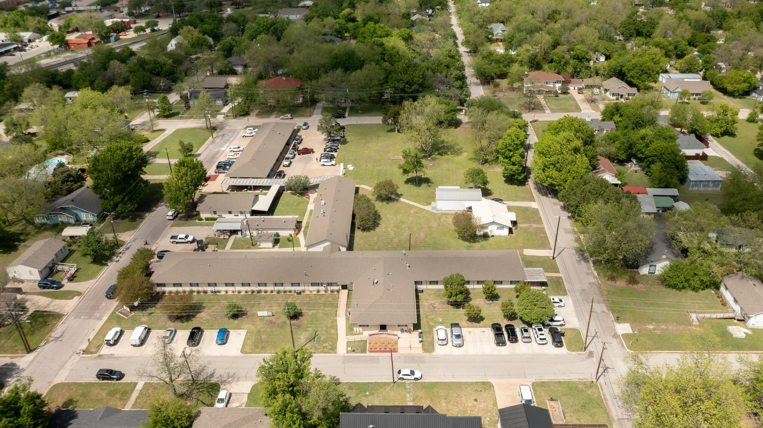 Aerial view of a neighborhood with single-story buildings, green lawns, trees, parked cars, and houses.